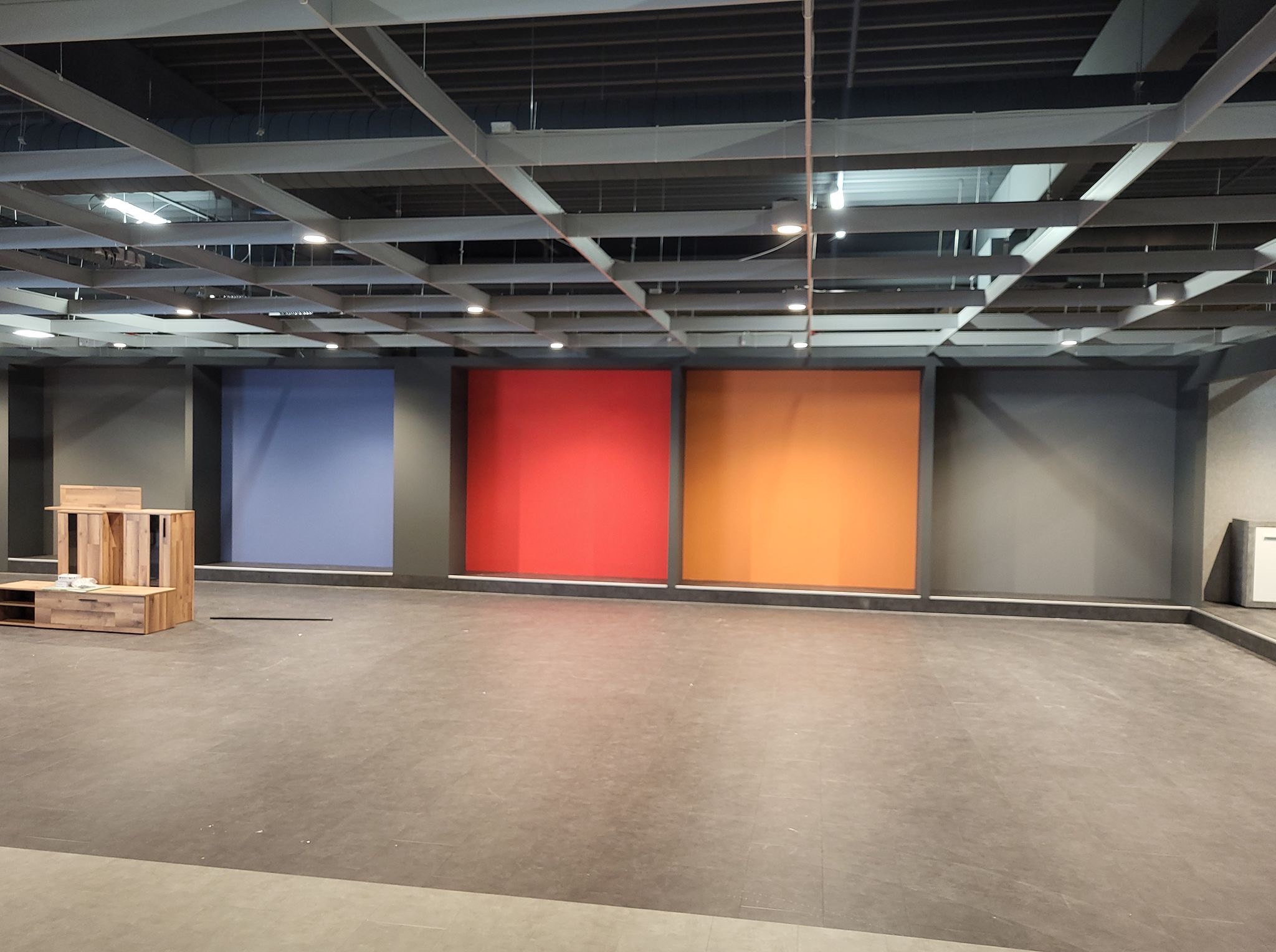 Wnętrze komercyjne z pomalowanymi na różne kolory ścianami (niebieski, czerwony, pomarańczowy). Widoczna szara podłoga i drewniane meble. Oświetlenie sufitowe.