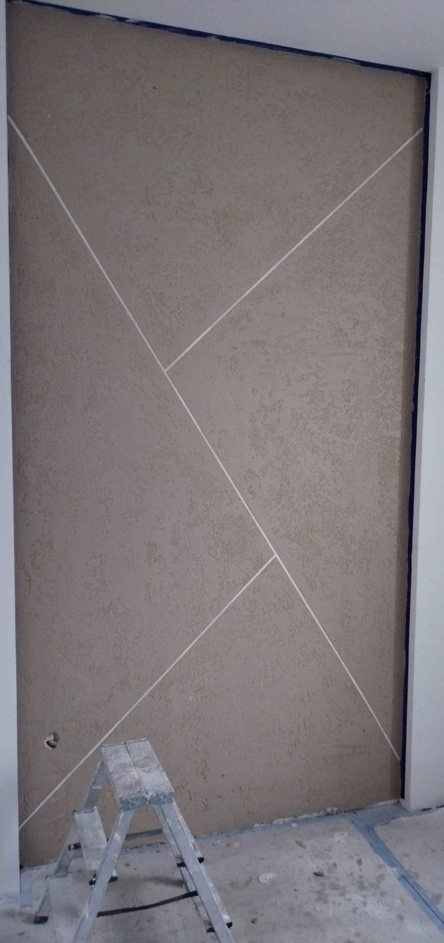 Dekoracyjna ściana z beżową strukturą i białymi, ukośnymi liniami. Widoczna drabina malarska i fragment podłogi. Oryginalne wykończenie wnętrza.