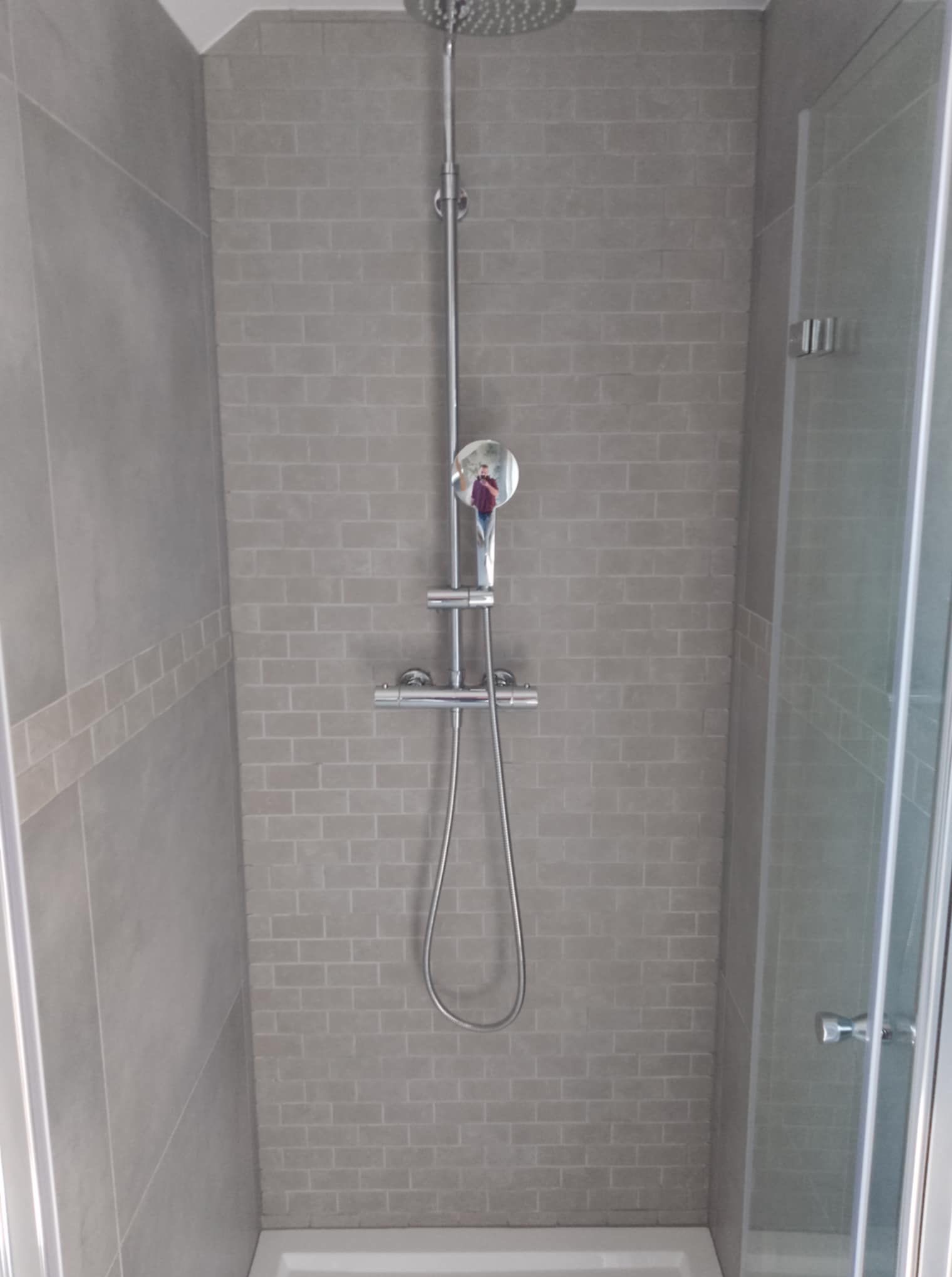 Nowoczesna kabina prysznicowa z szarymi płytkami imitującymi cegłę i chromowaną armaturą. Widoczna szklana ściana kabiny i deszczownica.