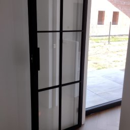 PirSpawArt Rafał Pawełek - Nowoczesne drzwi wewnętrzne z czarną ramą i szklanymi panelami, widoczne z bliska na tle jasnej ściany i podłogi w jodełkę, z widokiem na niedokończony budynek przez otwarte drzwi.