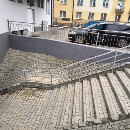PirSpawArt Rafał Pawełek - Zewnętrzne schody z szarej kostki brukowej prowadzące w dół, zabezpieczone metalową, srebrną balustradą, z widocznym fragmentem parkingu i samochodem osobowym w tle.