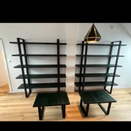 Dwa minimalistyczne, ciemnozielone stoliki kawowe i dwie otwarte, metalowe półki na tle białej ściany i drewnianej podłogi, z dekoracyjną lampą na jednej z półek.