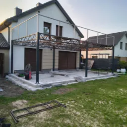 Konstrukcja metalowa pergoli przy domu w trakcie montażu, widoczny spawacz i elementy konstrukcyjne na trawniku. Ujęcie z dołu, w tle budynek i błękitne niebo.