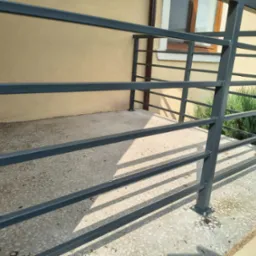 Szara, pozioma balustrada metalowa z profili zamkniętych na tarasie domu jednorodzinnego. Widoczne detale mocowania do podłoża i cień rzucany przez konstrukcję.