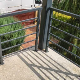 Nowoczesna, szara balustrada metalowa z poziomymi szczeblami na balkonie z widokiem na zieleń i brukowaną ścieżkę. Cień balustrady na podłodze balkonu.