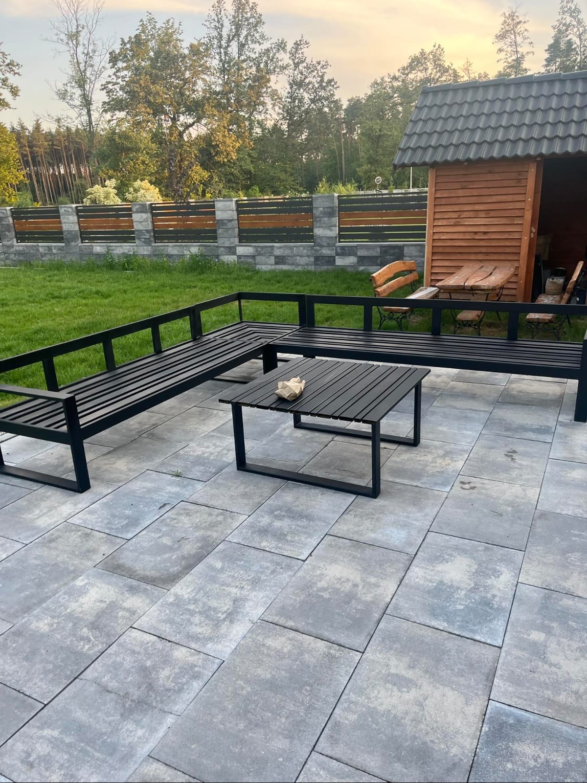 Nowoczesny zestaw mebli ogrodowych z czarnego metalu na szarym brukowanym patio, z widocznym ogrodzeniem z elementami drewnianymi i murowanymi w tle, oraz drewniana altana z meblami.