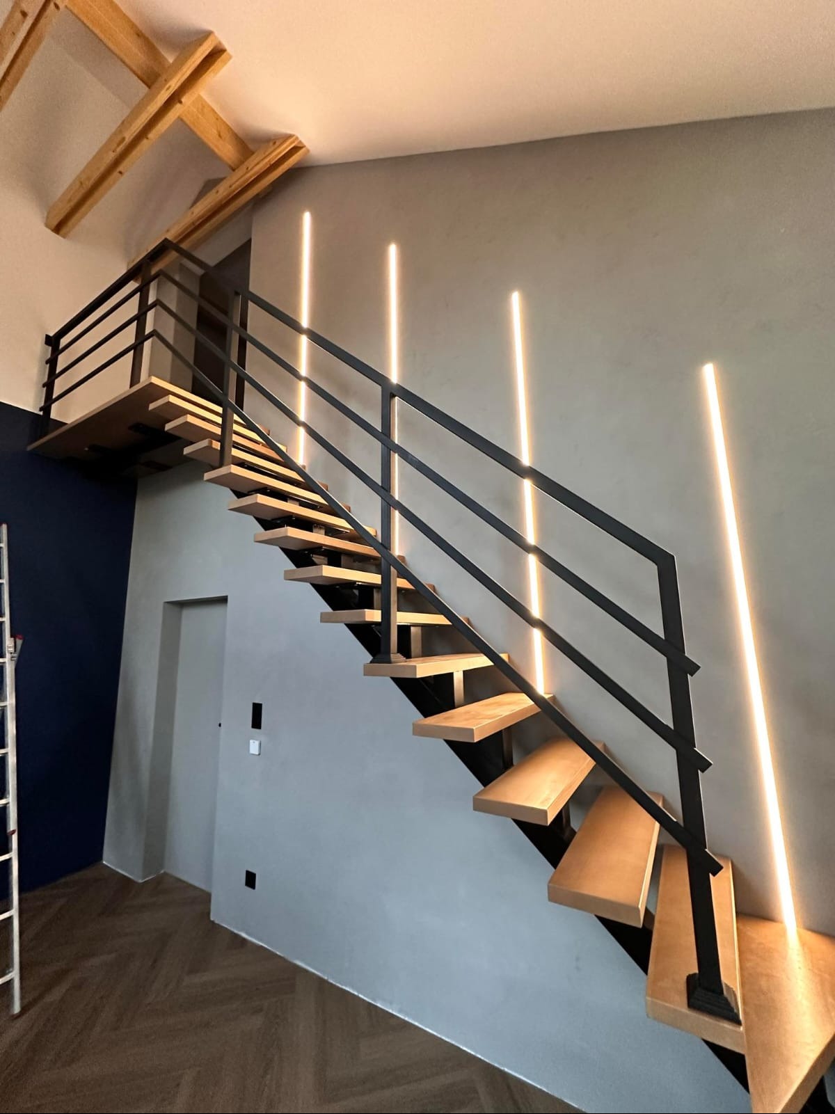 Nowoczesne schody metalowe z drewnianymi stopniami, podświetlone pionowymi liniami LED na szarej ścianie, widok z dołu na górę.