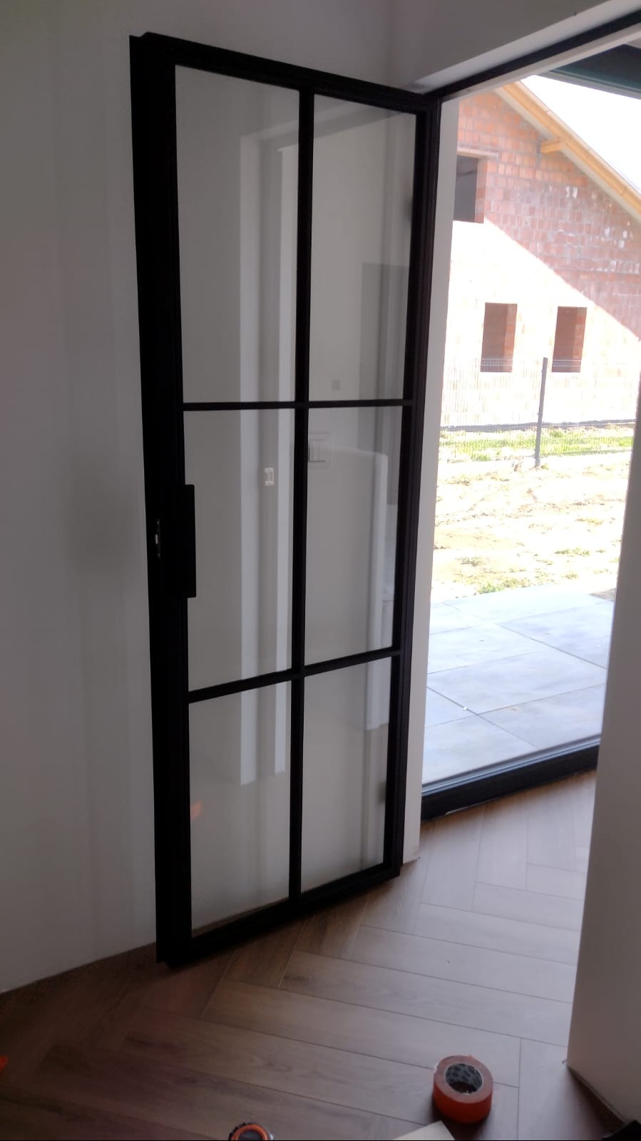 Nowoczesne drzwi wewnętrzne z czarną ramą i szklanymi panelami, widoczne z bliska na tle jasnej ściany i podłogi w jodełkę, z widokiem na niedokończony budynek przez otwarte drzwi.