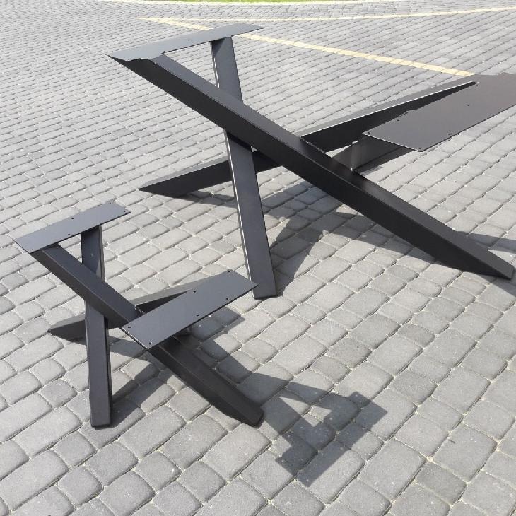 Dwie czarne, metalowe podstawy stołów o geometrycznym kształcie, ustawione na szarej kostce brukowej w słoneczny dzień. Większa podstawa ma kształt litery X z dodatkowymi elementami mocującymi,...