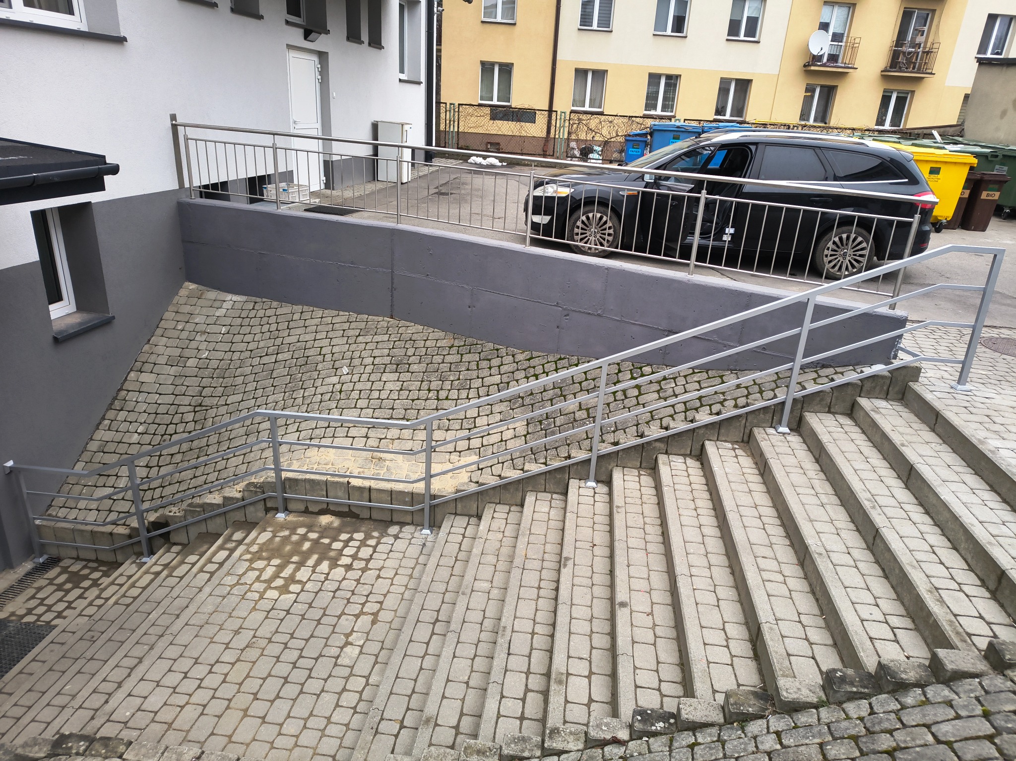 Zewnętrzne schody z szarej kostki brukowej prowadzące w dół, zabezpieczone metalową, srebrną balustradą, z widocznym fragmentem parkingu i samochodem osobowym w tle.