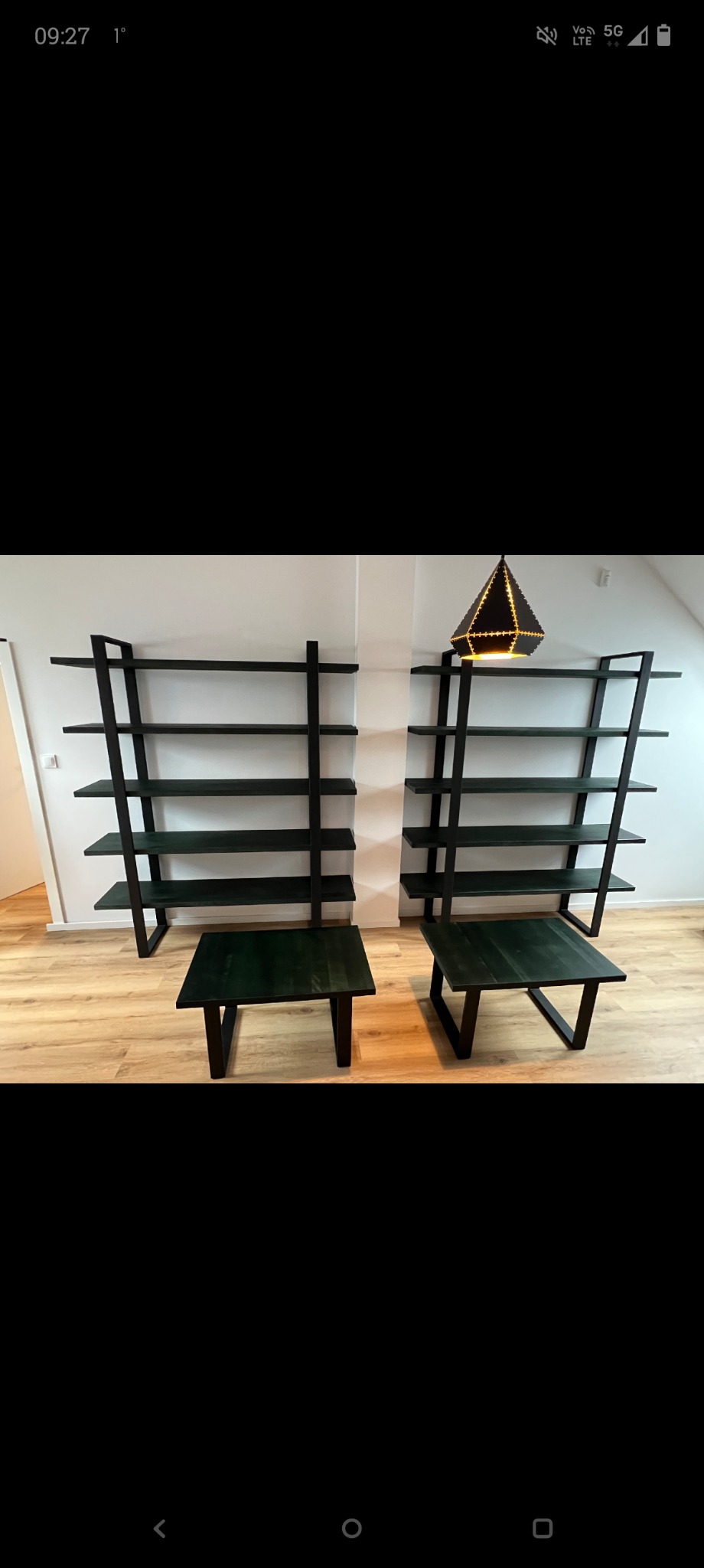 Dwa minimalistyczne, ciemnozielone stoliki kawowe i dwie otwarte, metalowe półki na tle białej ściany i drewnianej podłogi, z dekoracyjną lampą na jednej z półek.