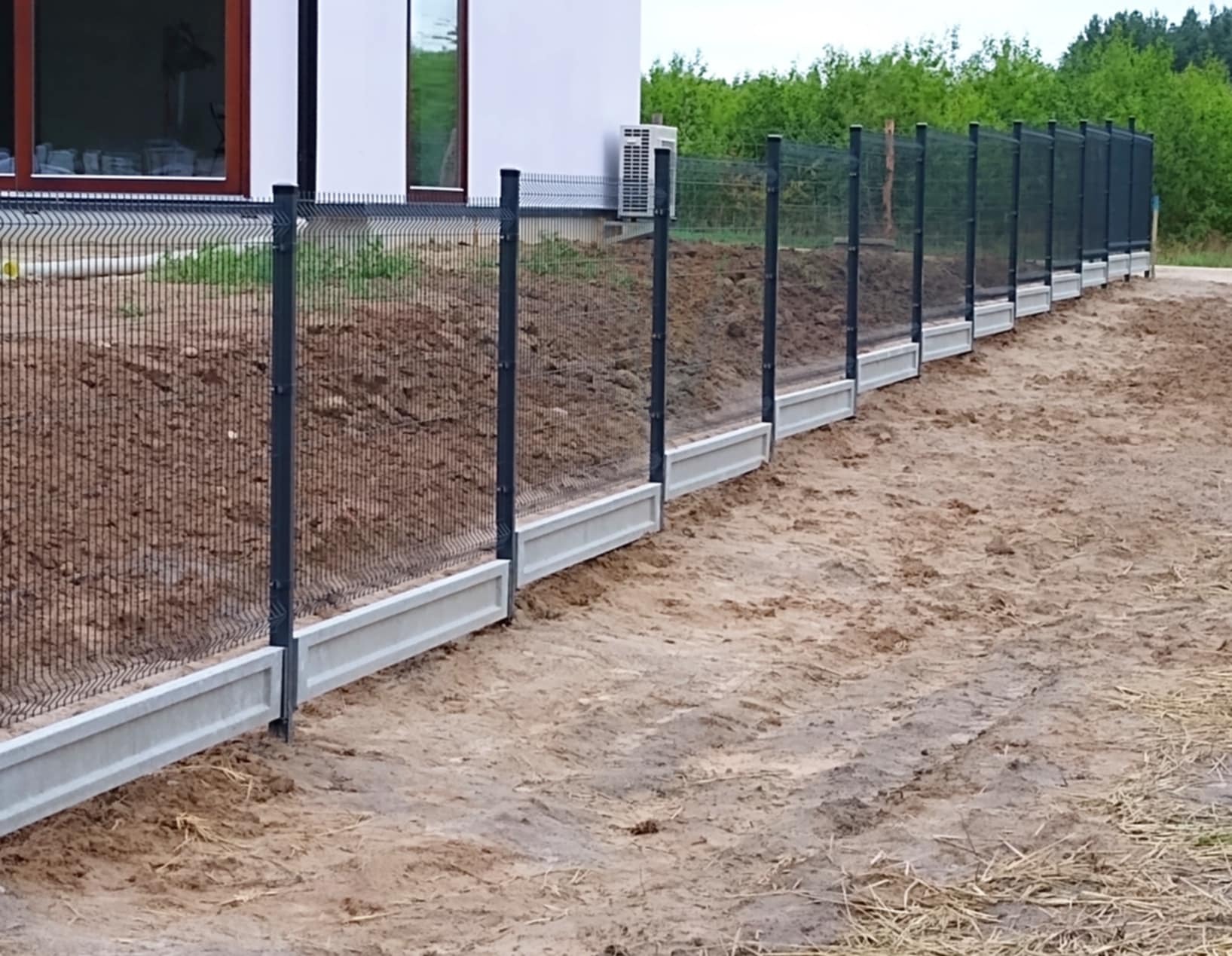 Nowoczesne ogrodzenie panelowe z podmurówką betonową, widok z zewnątrz, częściowo zasypane ziemią, z klimatyzatorem na ścianie budynku w tle.