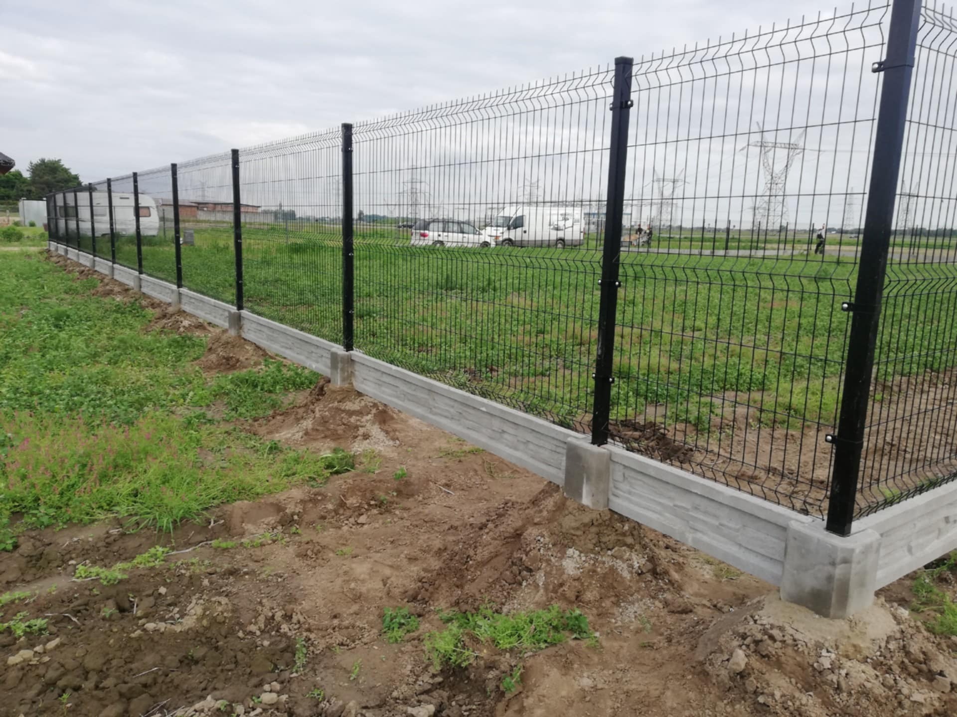 Nowo zainstalowane ogrodzenie panelowe z czarnymi słupkami i betonową podmurówką na nierównym terenie, w tle widoczne domy i słupy wysokiego napięcia.