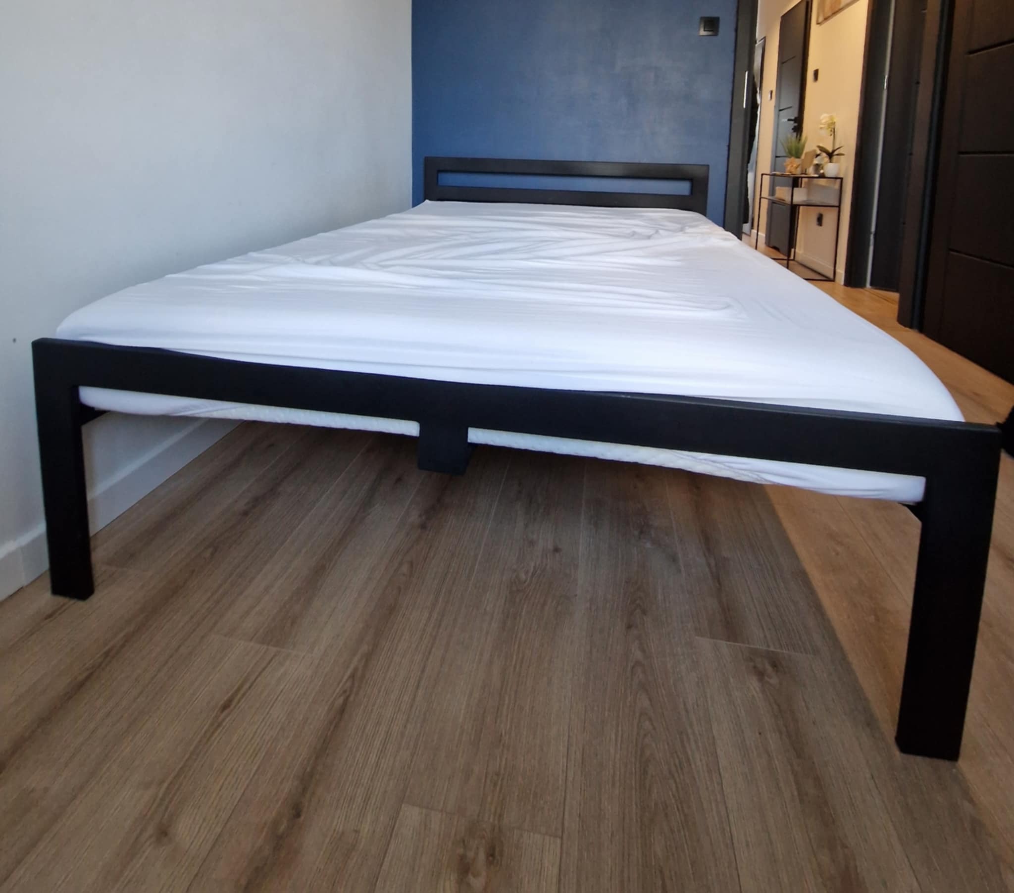 Metalowa rama łóżka z białym prześcieradłem na drewnianej podłodze, widok z dołu, w tle niebieska ściana i elementy wyposażenia wnętrza.