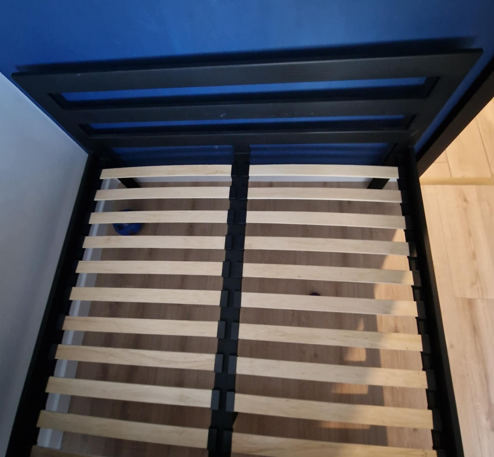 Rama łóżka z drewnianym stelażem, czarna metalowa konstrukcja na tle niebieskiej ściany, widok z góry na podłogę.