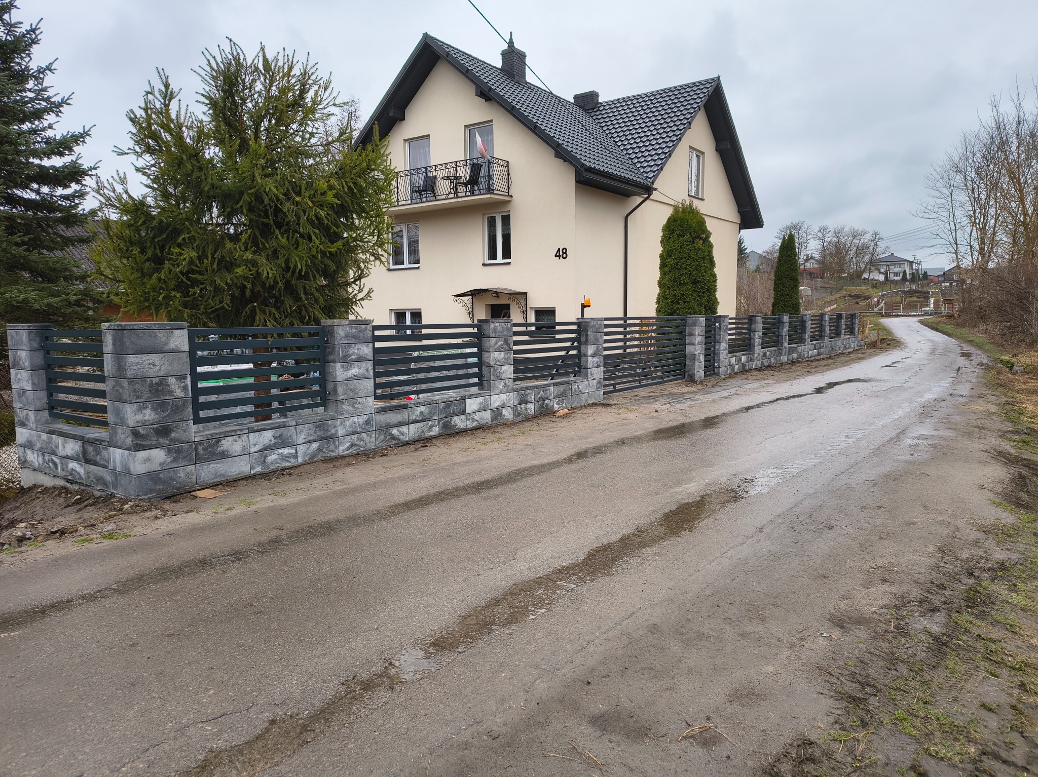 Nowoczesne ogrodzenie panelowe z betonową podmurówką otaczające dom z balkonem, widok z zewnątrz na mokrą drogę dojazdową.