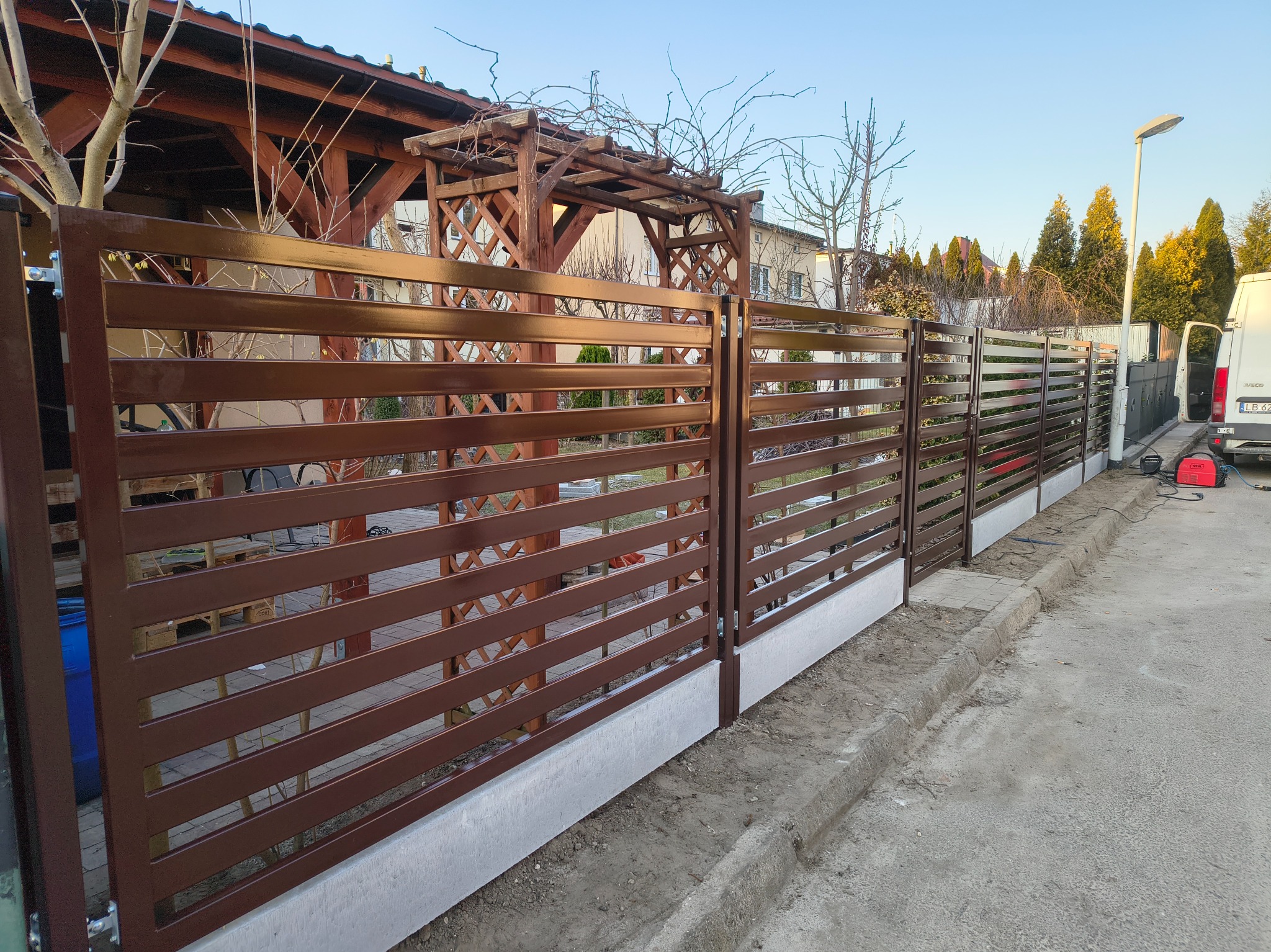 Nowoczesne, poziome ogrodzenie panelowe w kolorze brązowym na niskim, betonowym fundamencie, z drewnianą pergolą w tle i białym samochodem dostawczym w pobliżu.