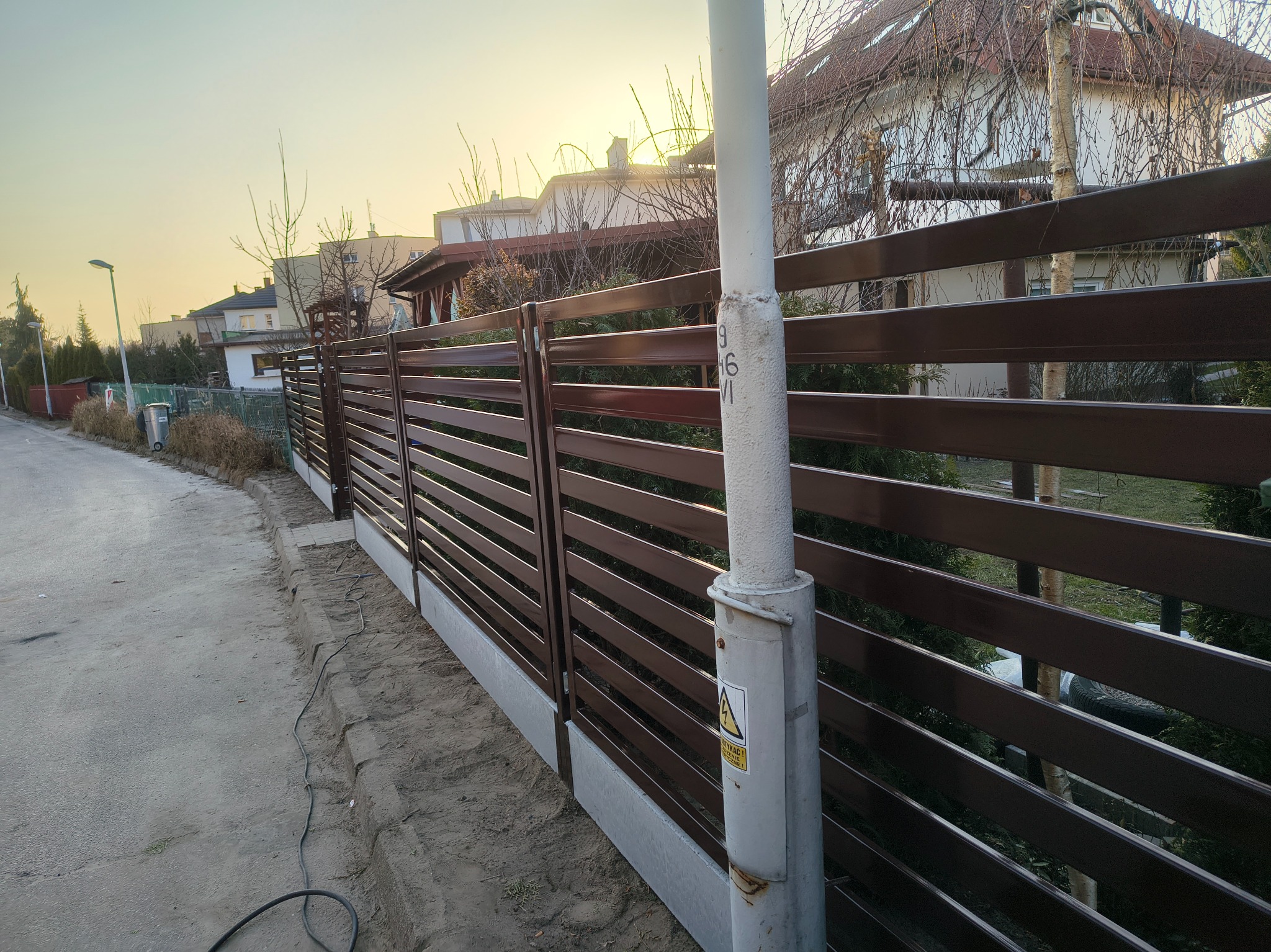 Nowoczesne, poziome ogrodzenie z brązowych paneli na betonowej podmurówce, widziane z perspektywy ulicy w słoneczny dzień. Słup energetyczny z naklejką ostrzegawczą w pierwszym planie.