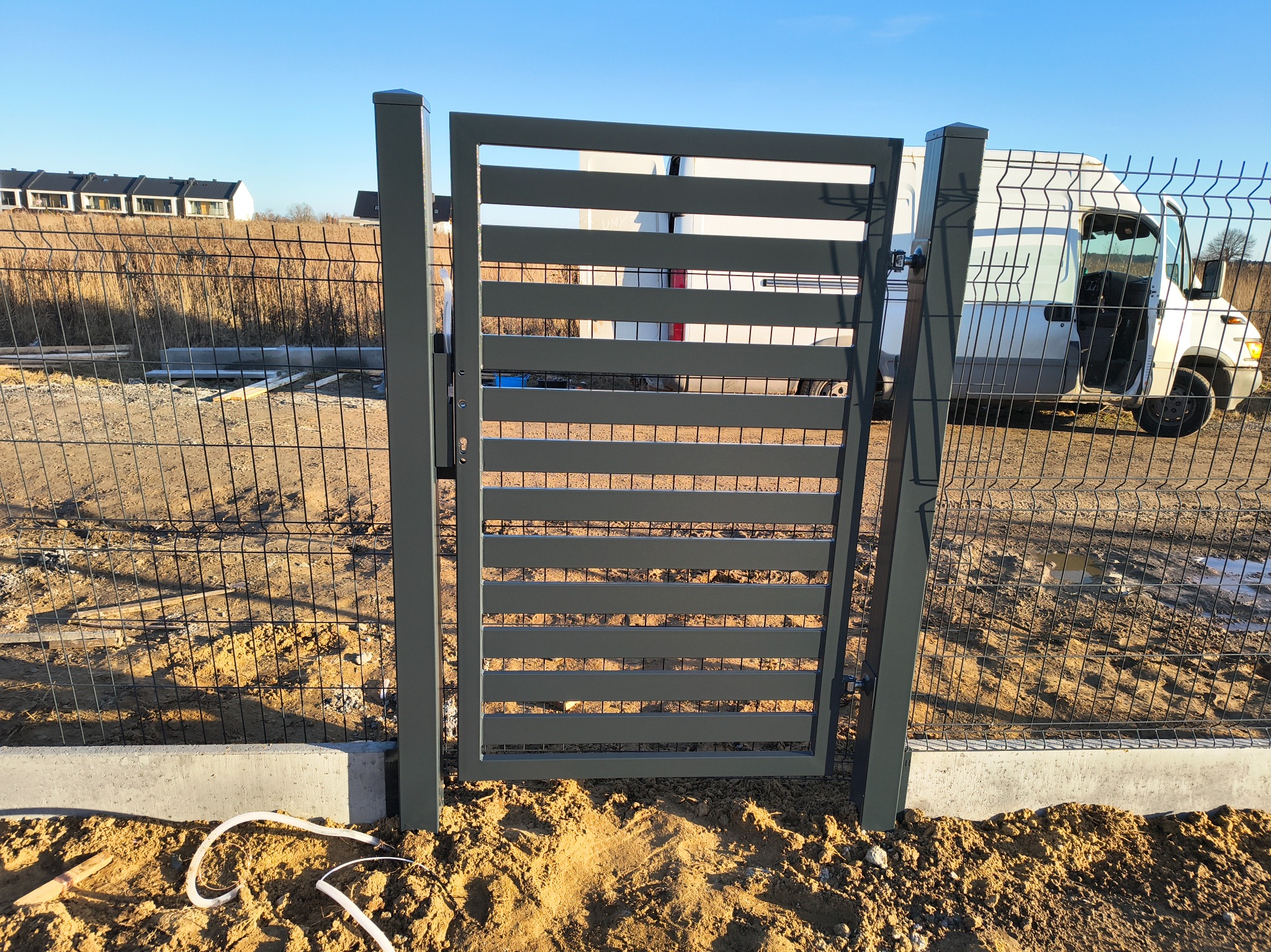 Nowoczesna, pozioma brama wjazdowa z metalowymi panelami, widoczna na tle ogrodzenia z siatki i białego samochodu dostawczego w słoneczny dzień.