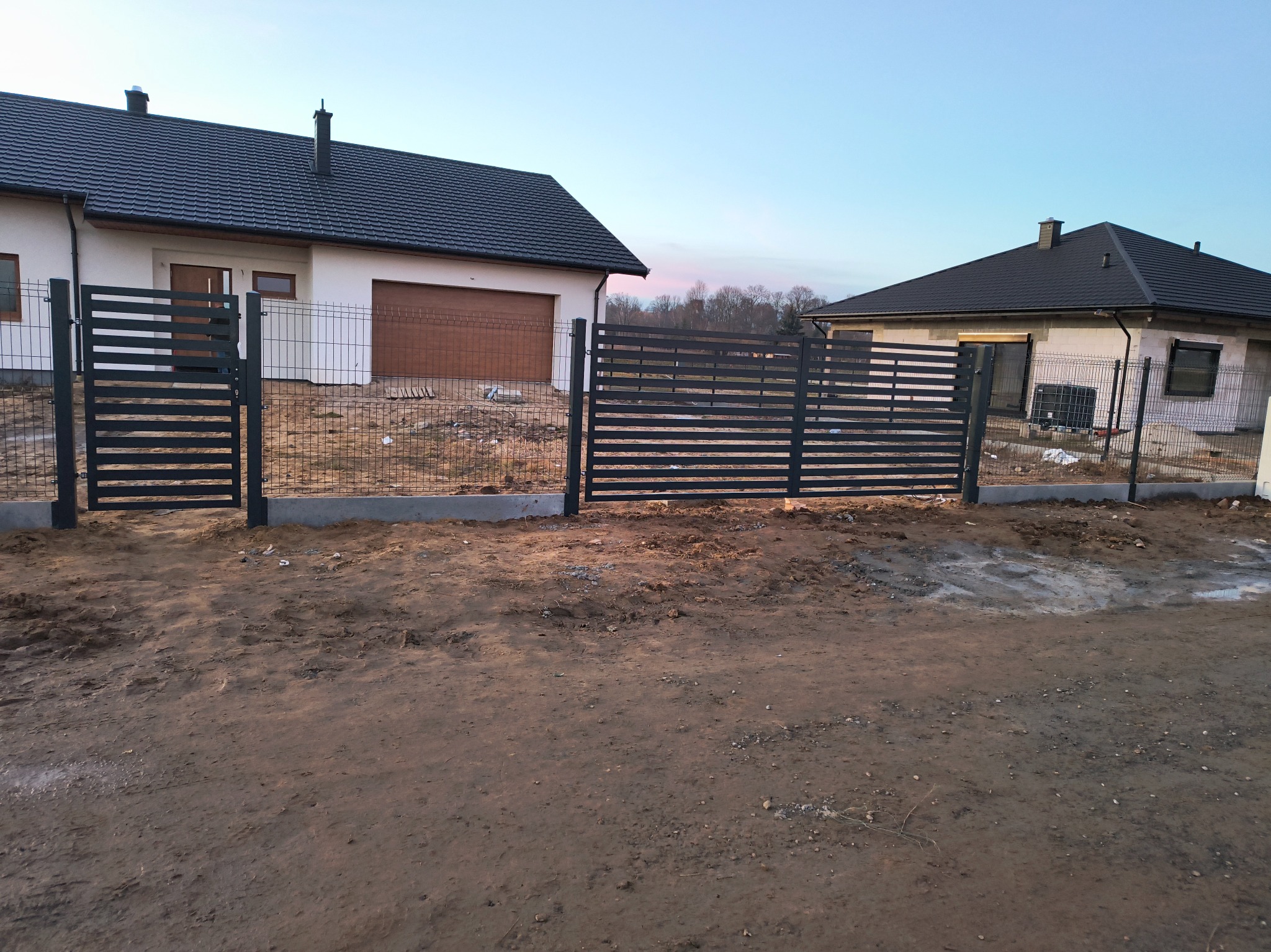 Nowoczesne, poziome przęsła ogrodzeniowe z metalu w kolorze grafitowym, oddzielające dwie posesje z nowymi domami jednorodzinnymi w trakcie wykańczania, widoczne świeże wylewki fundamentów...