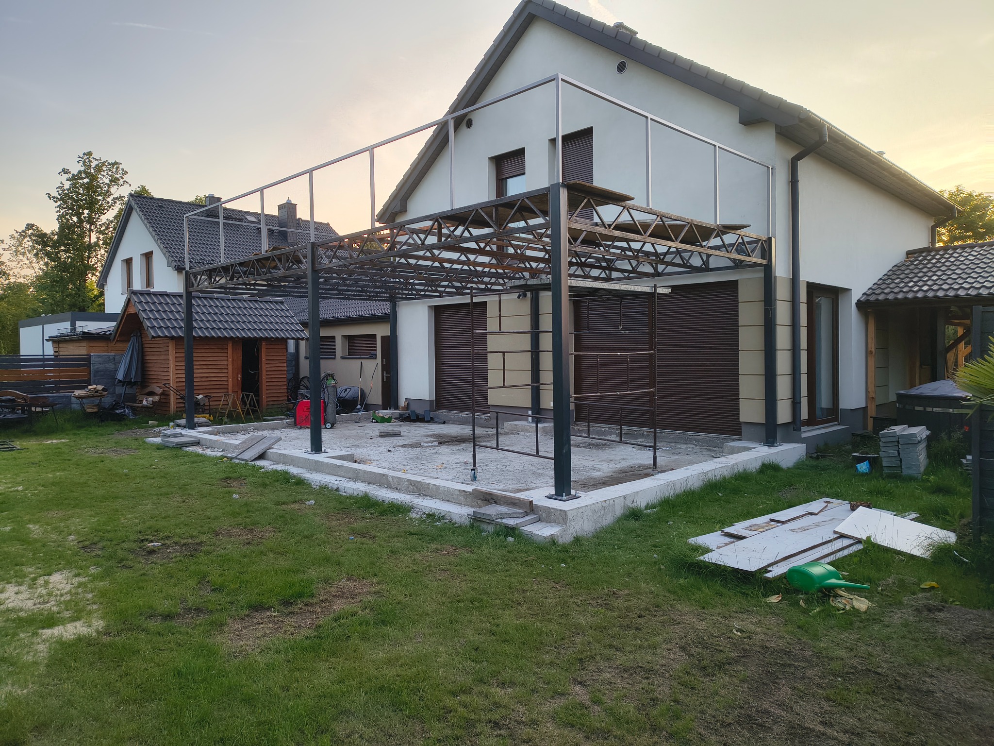 Metalowa konstrukcja pergoli w trakcie montażu na betonowym fundamencie, widok z zewnątrz, dom w tle.