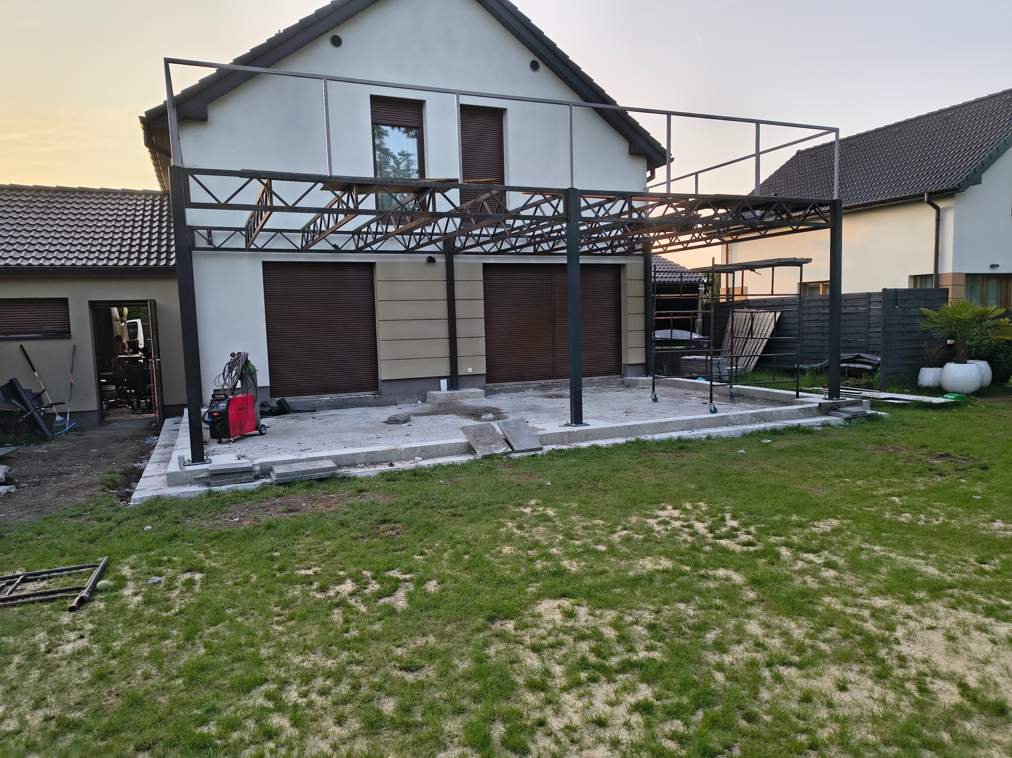 W trakcie budowy metalowej pergoli przy domu jednorodzinnym. Widoczny szkielet konstrukcji, fundament i sprzęt spawalniczy. Tło stanowi budynek z brązowymi roletami.