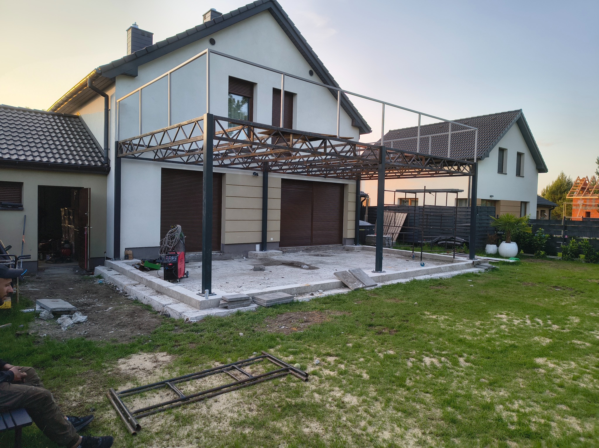 Konstrukcja metalowa pergoli przy domu w trakcie montażu, widoczny spawacz i elementy konstrukcyjne na trawniku. Ujęcie z dołu, w tle budynek i błękitne niebo.