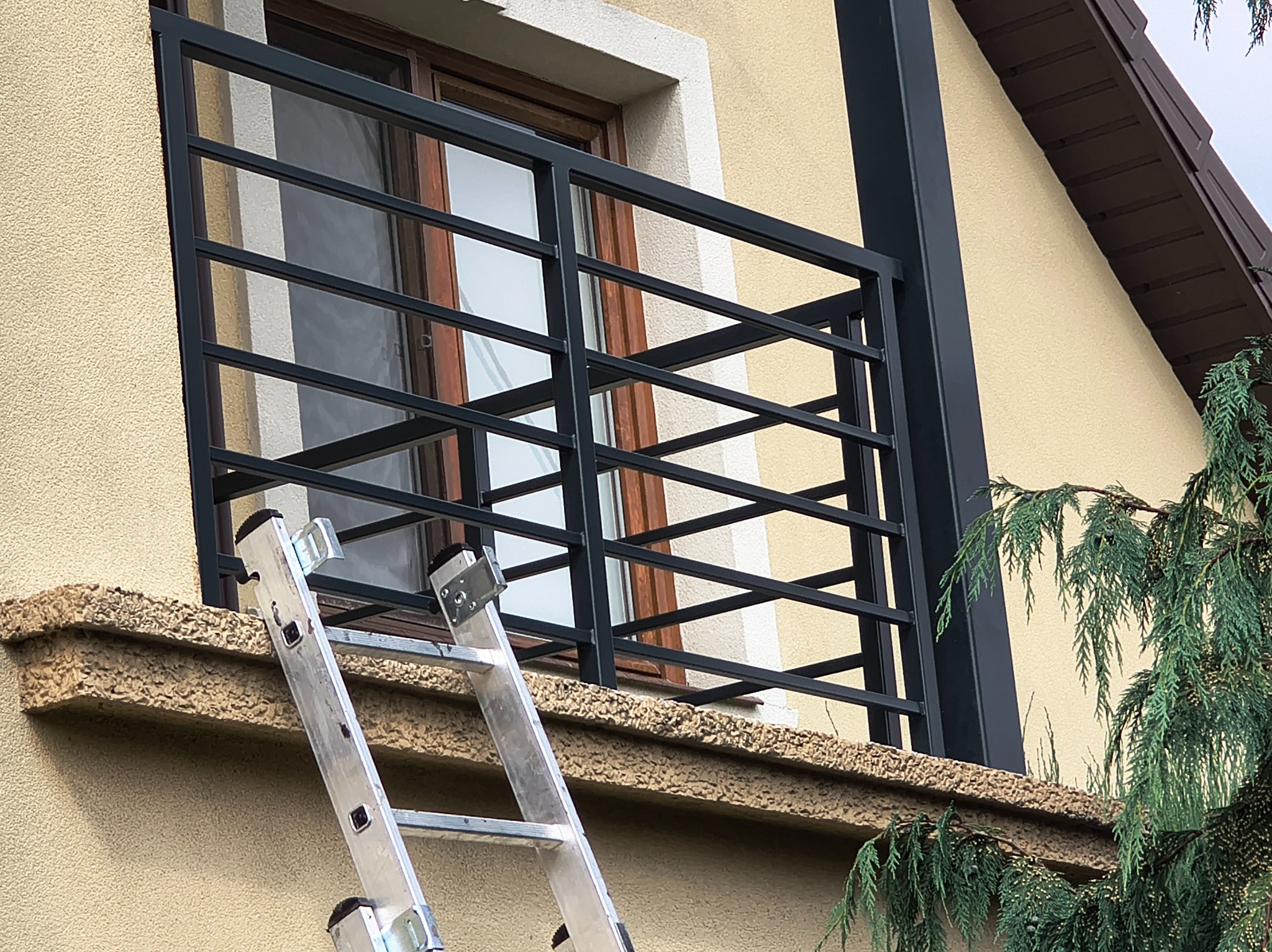 Czarna, pozioma balustrada balkonowa z metalu, widoczna z dołu, z drabiną opartą o balkon. Tło stanowi beżowa elewacja budynku i fragment zieleni.