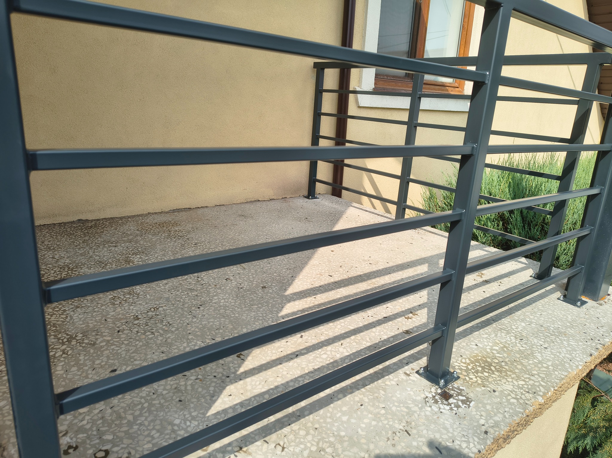 Szara, pozioma balustrada metalowa z profili zamkniętych na tarasie domu jednorodzinnego. Widoczne detale mocowania do podłoża i cień rzucany przez konstrukcję.