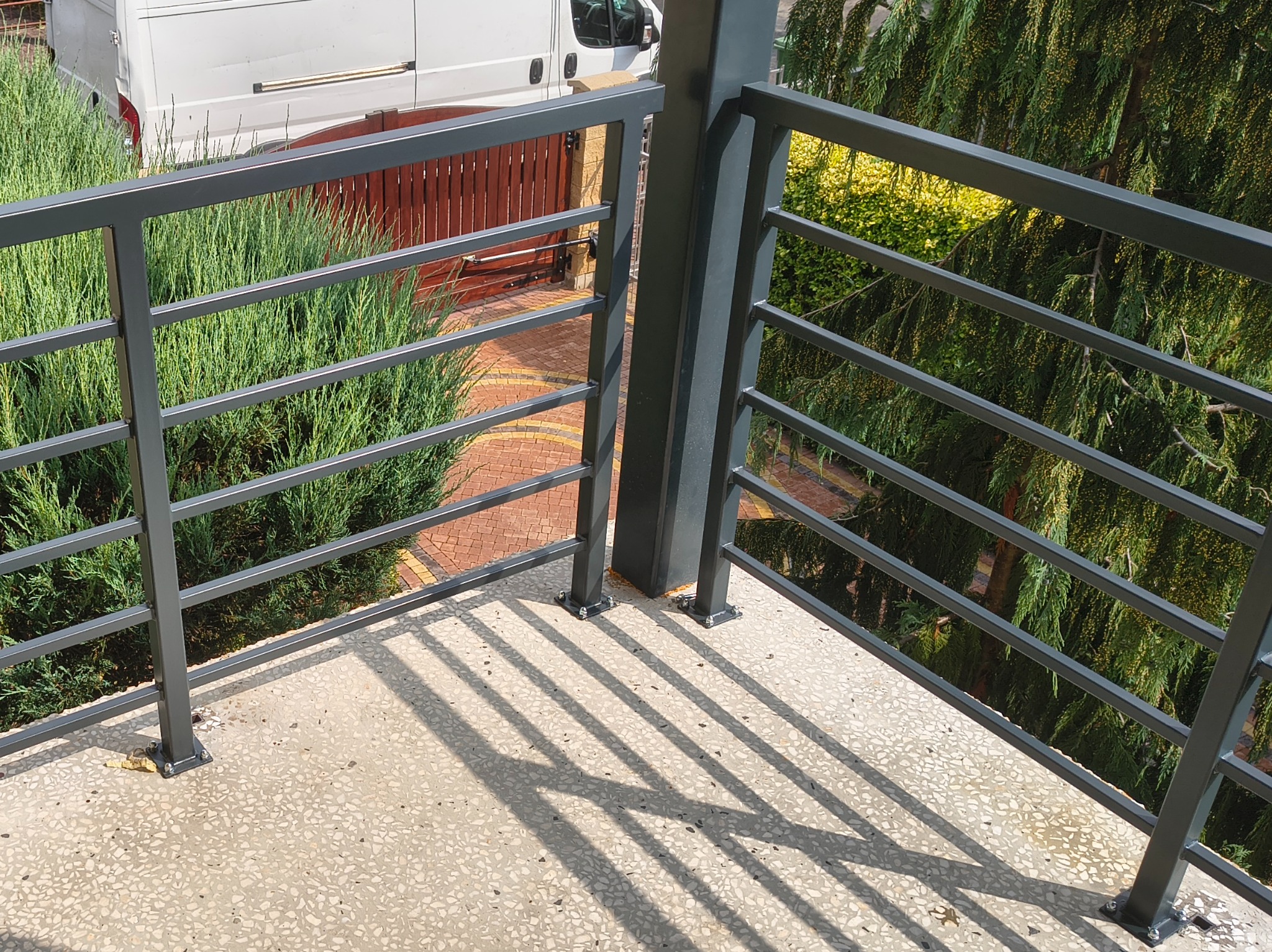 Nowoczesna, szara balustrada metalowa z poziomymi szczeblami na balkonie z widokiem na zieleń i brukowaną ścieżkę. Cień balustrady na podłodze balkonu.