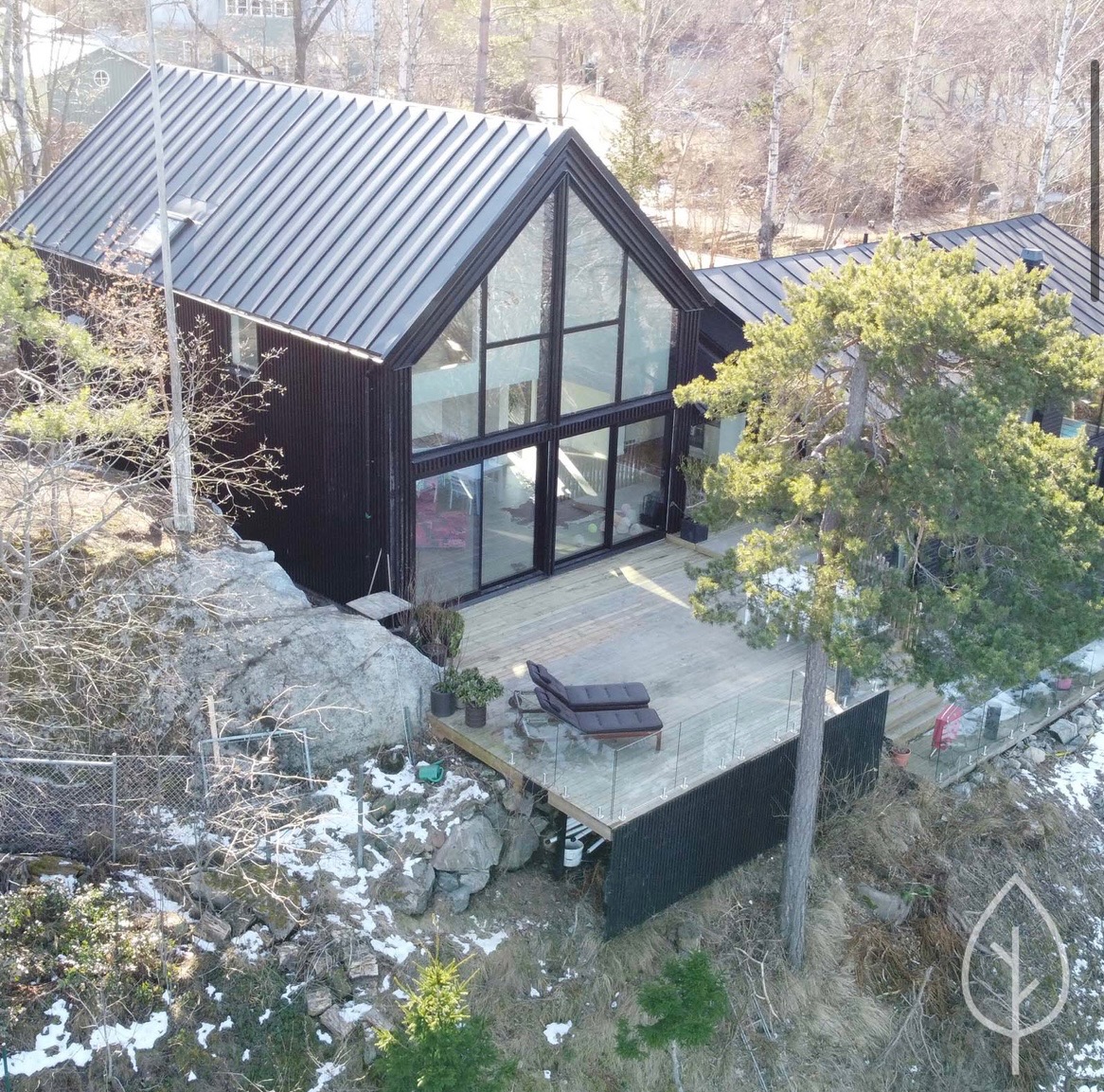 Nowoczesny dom z czarną elewacją i dużymi przeszkleniami, drewniany taras z leżakami, otoczony naturalnym krajobrazem ze skałami i drzewami.