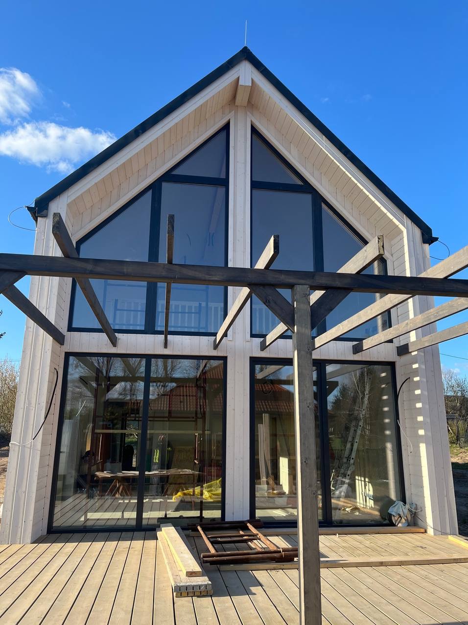 Surowy stan domu w Skrzeszewie z dużymi oknami i drewnianym tarasem, widoczne elementy konstrukcyjne pergoli.