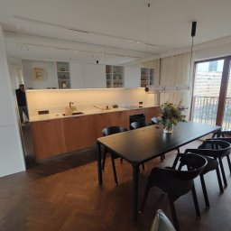 Dwa Fronty - Nowoczesna kuchnia z drewnianymi frontami szafek, białymi szafkami wiszącymi i blatem z jasnego kamienia, oświetlona listwą LED pod szafkami, widok na stół z czarnymi krzesłami i bukiet kwiatów, za...