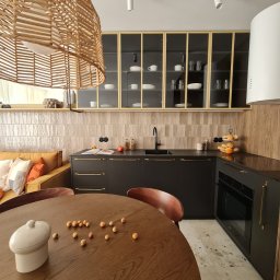 Dwa Fronty - Nowoczesna kuchnia we Wrocławiu z czarnymi szafkami i złotymi uchwytami, przeszklonymi szafkami wiszącymi z widoczną zastawą oraz okrągłym drewnianym stołem z dekoracjami.
