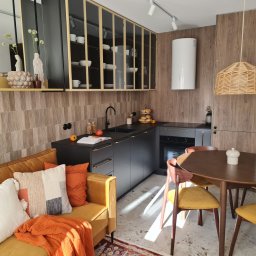 Dwa Fronty - Nowoczesna kuchnia z czarnymi szafkami, drewnianymi akcentami i żółtą sofą w salonie, oświetlona naturalnym światłem, z widocznym stołem jadalnym i dekoracjami.