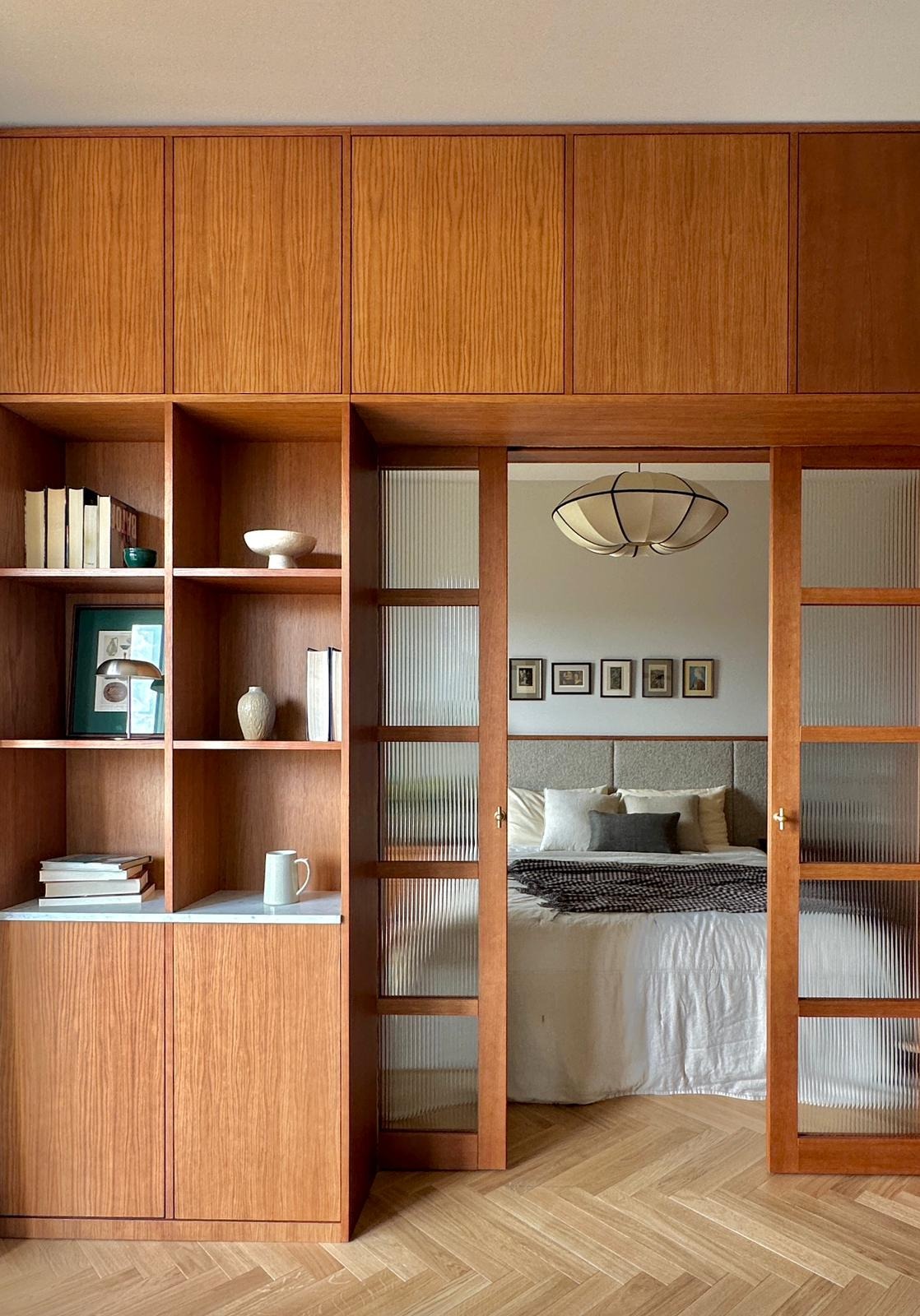 Drewniana zabudowa w sypialni z przesuwnymi drzwiami z ryflowanego szkła, regał z książkami i dekoracjami, parkiet w jodełkę.