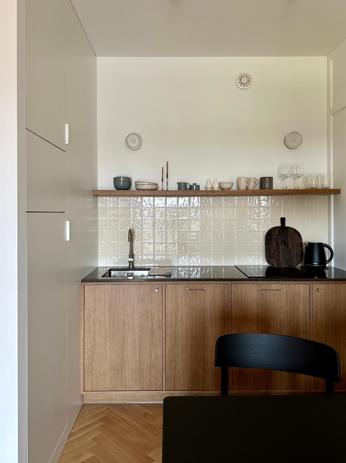 Nowoczesna kuchnia z drewnianymi frontami szafek, czarnym blatem i białymi, kwadratowymi kafelkami na ścianie. Na półce nad blatem ustawione dekoracje i naczynia.