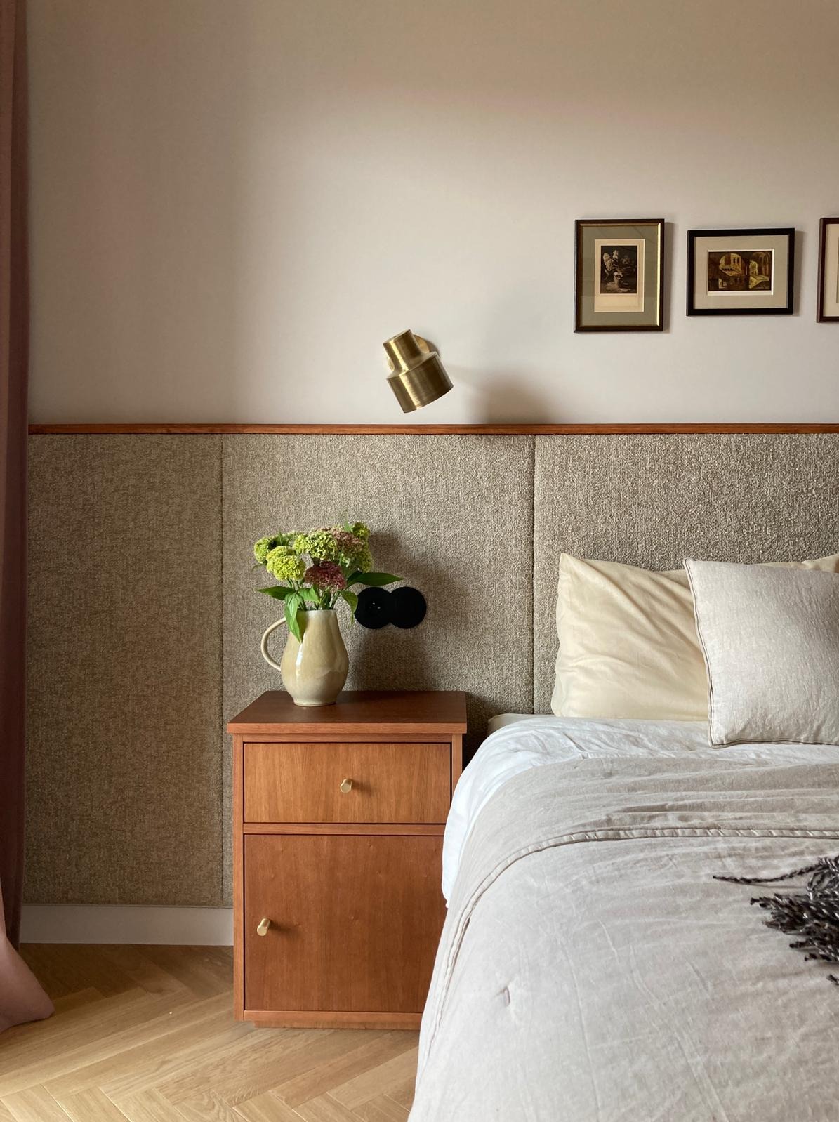 Nowoczesna sypialnia z drewnianą szafką nocną, na której stoi wazon z kwiatami. Nad szafką zamontowany jest kinkiet w kolorze mosiądzu. Wezgłowie łóżka obite jest tkaniną w neutralnym odcieniu.