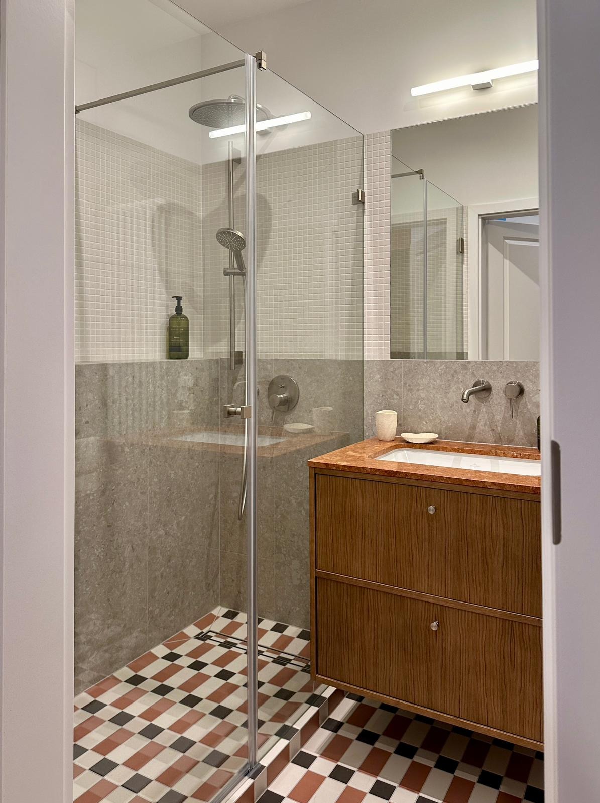 Nowoczesna łazienka z kabiną prysznicową ze szklanymi drzwiami, marmurową umywalką z drewnianą szafką i wzorzystą podłogą w kratkę.