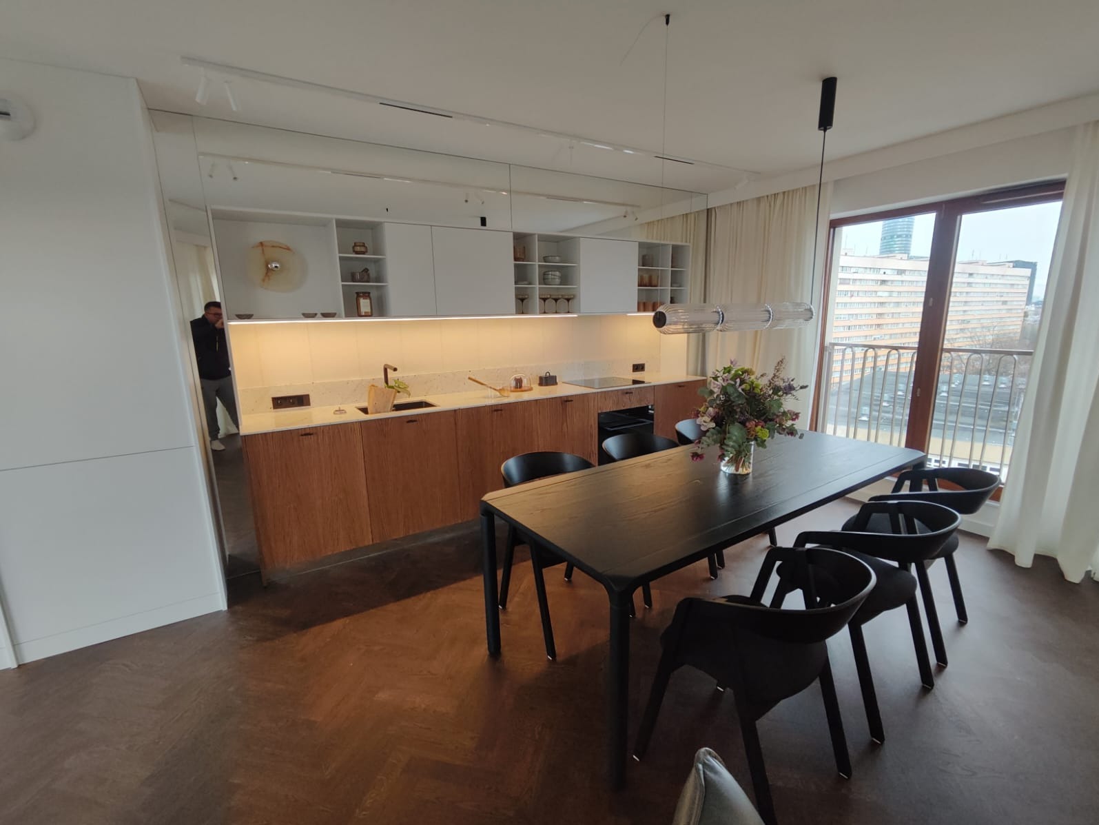 Nowoczesna kuchnia z drewnianymi frontami szafek, białymi szafkami wiszącymi i blatem z jasnego kamienia, oświetlona listwą LED pod szafkami, widok na stół z czarnymi krzesłami i bukiet kwiatów, za...