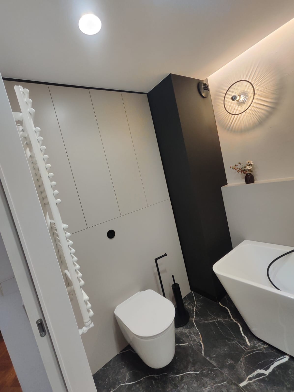 Nowoczesna łazienka w odcieniach szarości i czerni z marmurową posadzką, zabudową meblową pod sufit, wiszącą toaletą i wolnostojącą wanną.