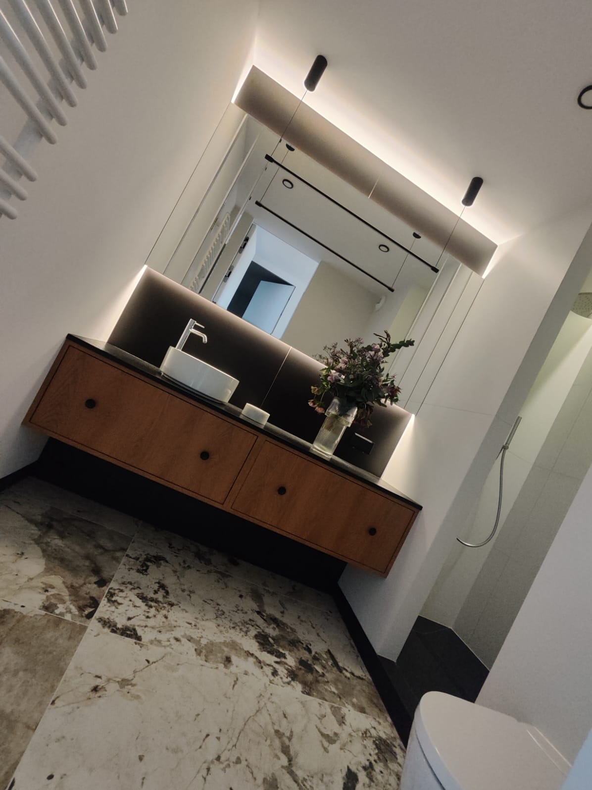 Nowoczesna łazienka z drewnianą szafką pod umywalką, marmurową podłogą i podświetlanym lustrem, uzupełniona bukietem kwiatów w szklanym wazonie.