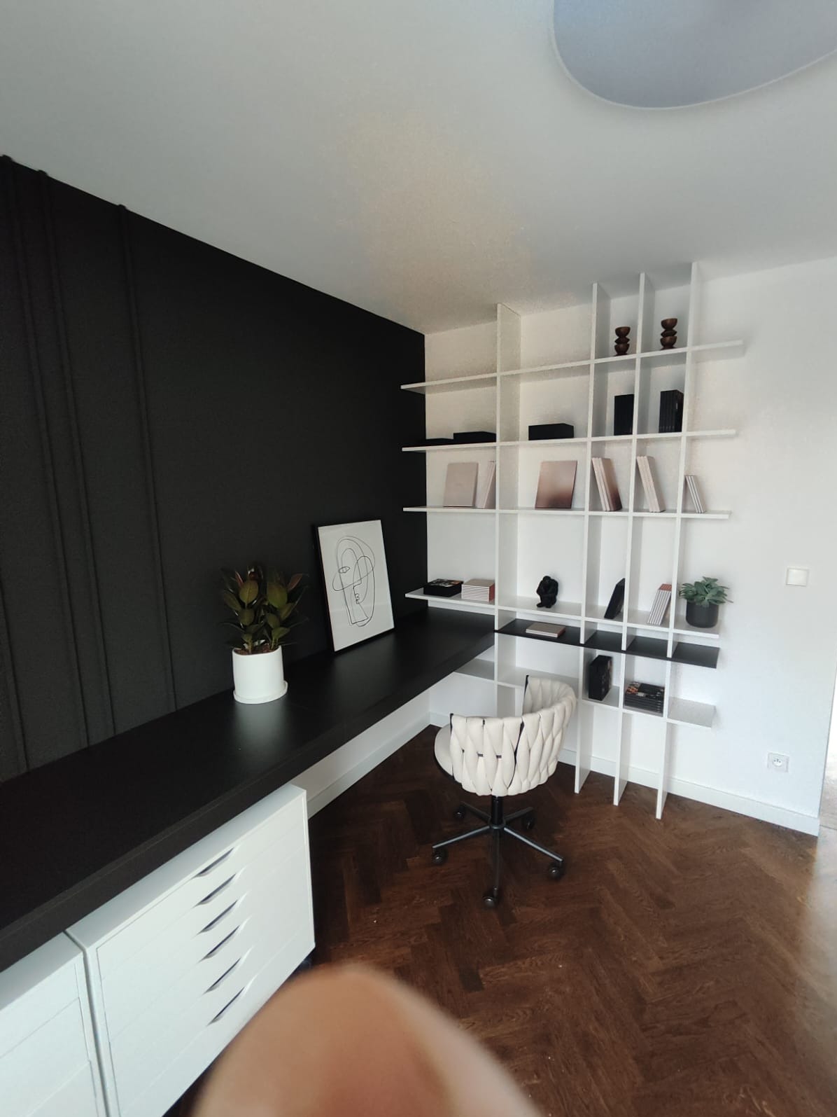 Nowoczesne biuro z czarnym blatem i białymi szafkami, uzupełnione białą, ażurową półką i krzesłem w stylu boho na parkiecie w jodełkę.