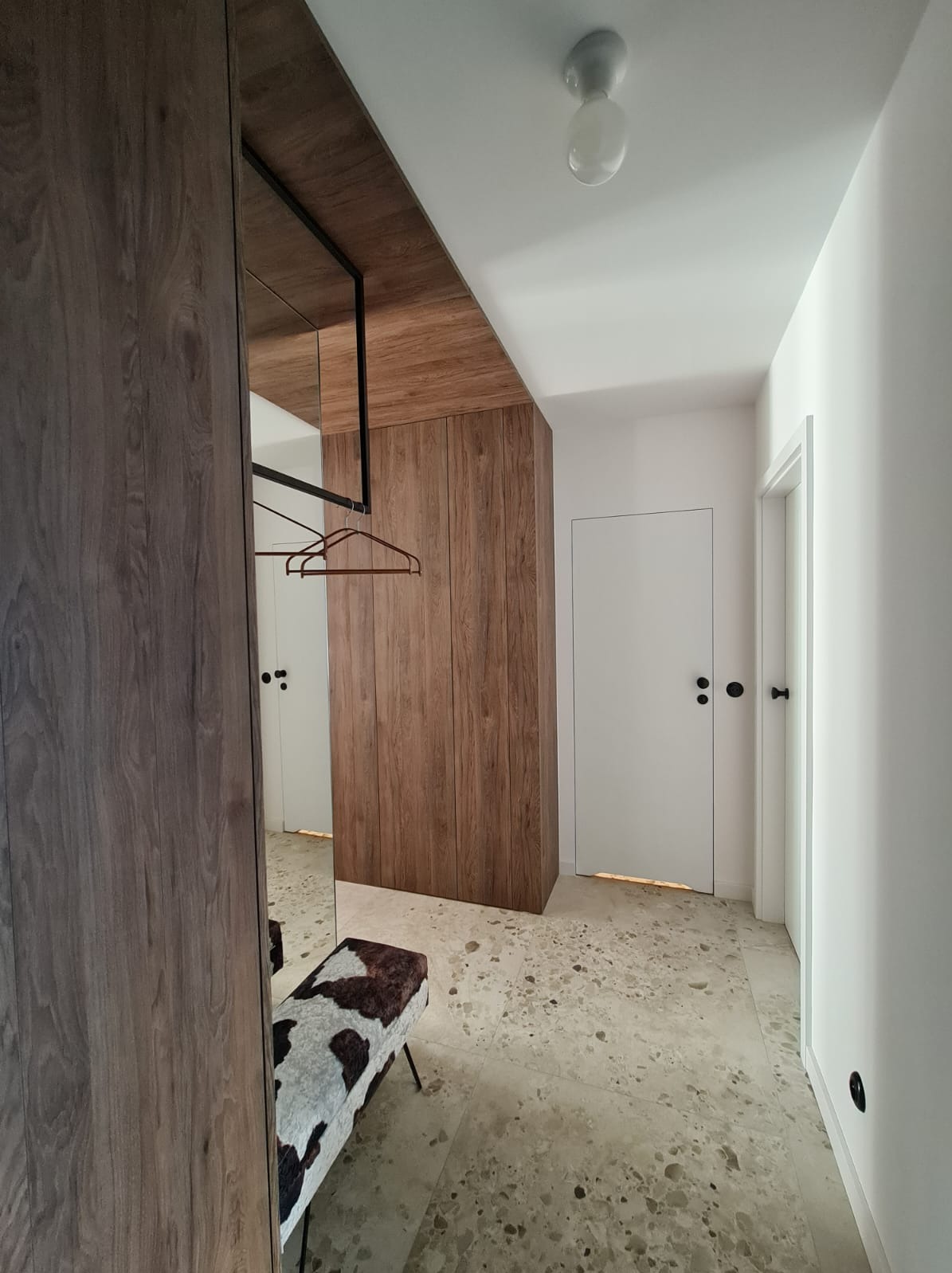 Nowoczesna szafa wnękowa z lustrem, drewnianym wykończeniem i siedziskiem w stylu krowiej skóry w przedpokoju z podłogą lastryko.