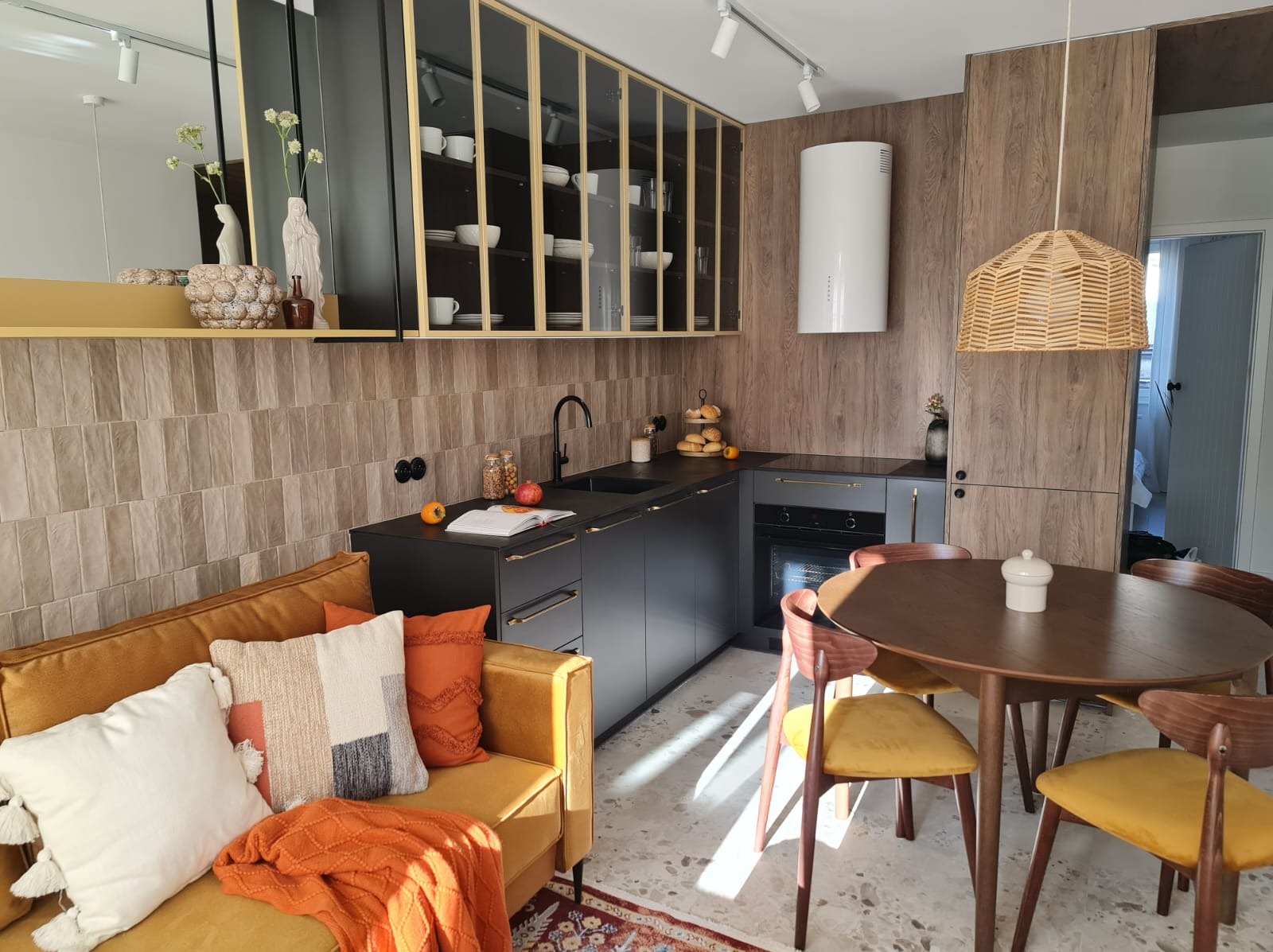 Nowoczesna kuchnia z czarnymi szafkami, drewnianymi akcentami i żółtą sofą w salonie, oświetlona naturalnym światłem, z widocznym stołem jadalnym i dekoracjami.