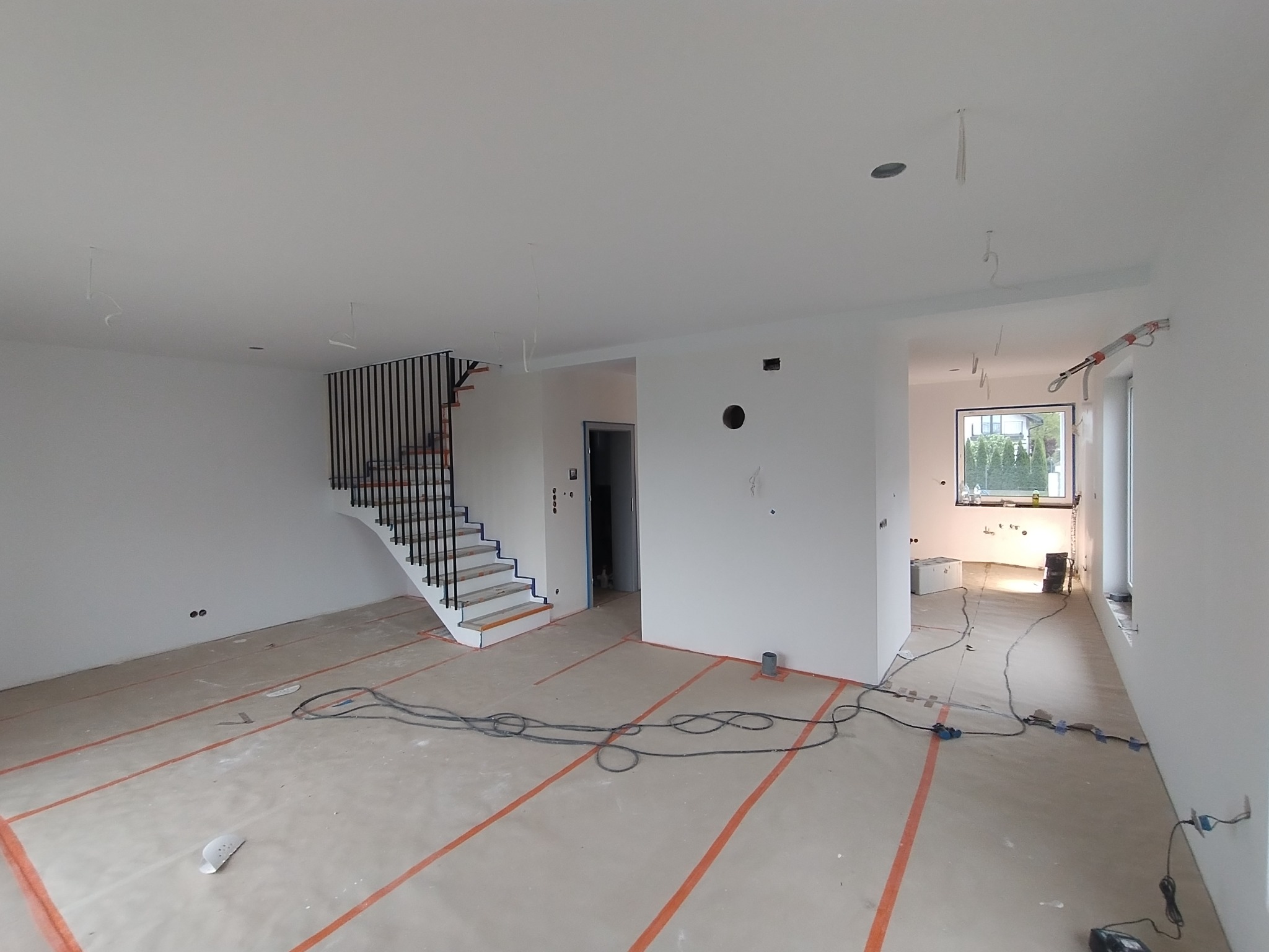 Surowe wnętrze domu w trakcie wykończenia: białe ściany, schody z czarną balustradą, przygotowane podłoże, widoczne kable i instalacje. Stan deweloperski.