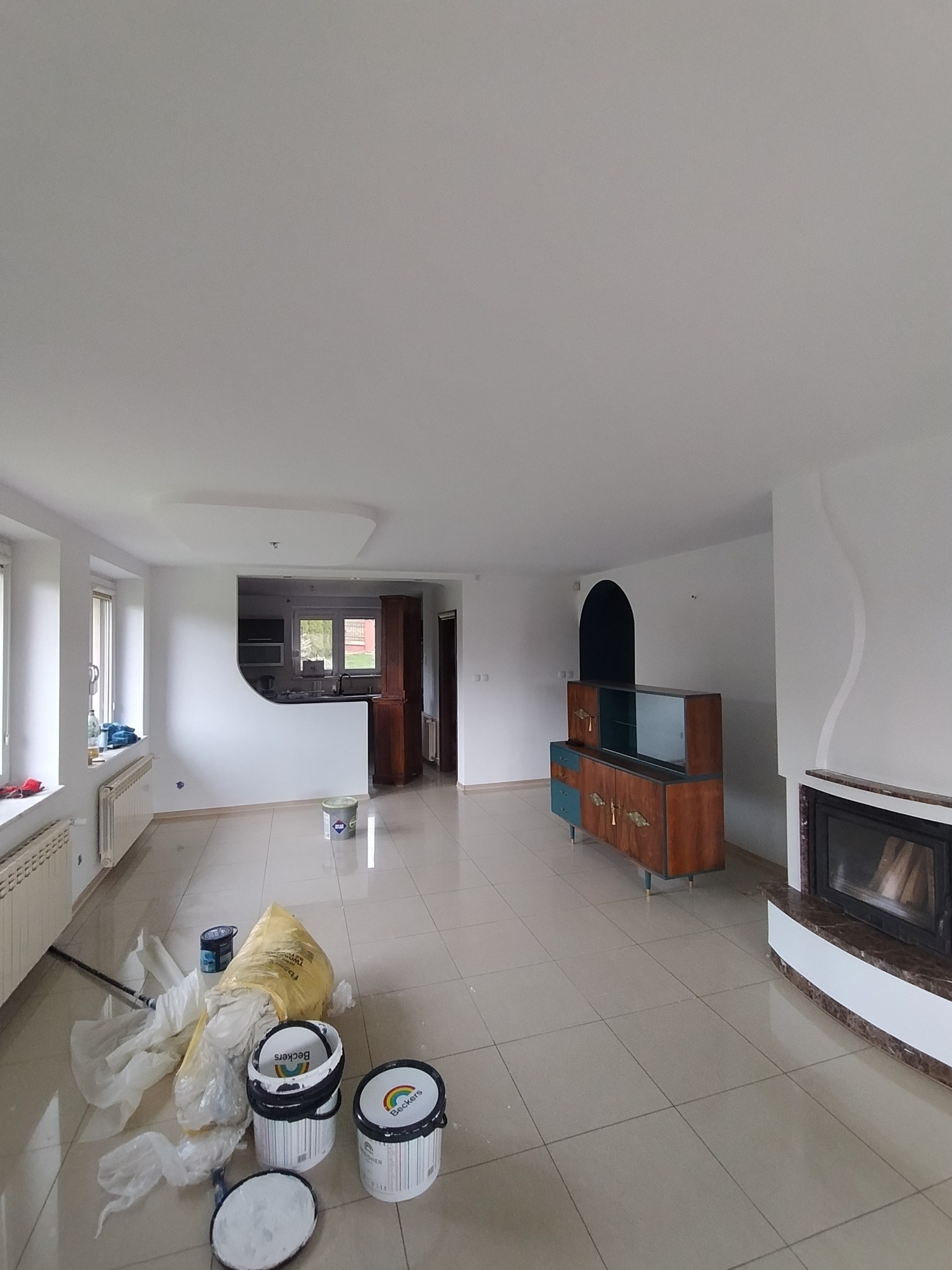 Jasne wnętrze po malowaniu ścian na biało, widoczne puszki z farbą, kominek i meble w stylu retro. Świeżo odmalowane ściany i czyste płytki podłogowe.