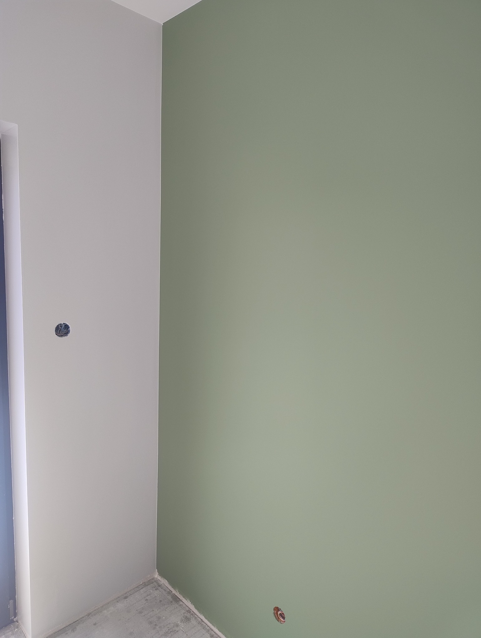 Pomieszczenie po malowaniu: ściana w kolorze zielonym i szara ściana z puszką elektryczną. Widoczny betonowy surowy podkład.