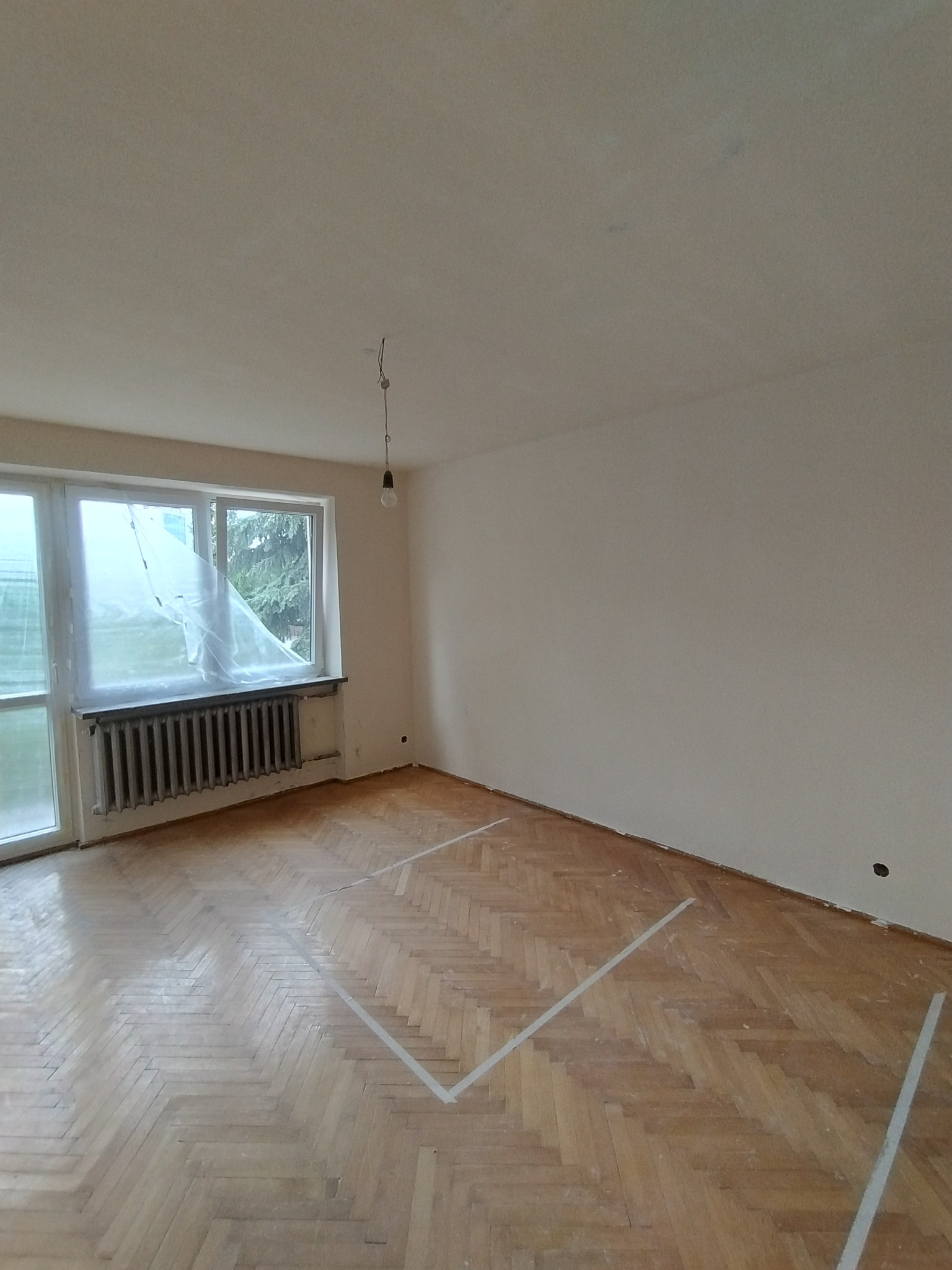 Pokój przed remontem: parkiet w jodełkę zabezpieczony taśmą malarską, ściany i sufit przygotowane do malowania, okno z zasłoniętym folią grzejnikiem.