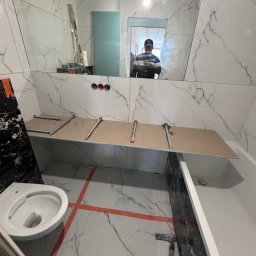 STROY BUD.group ANDRII PYLYPENKO - Łazienka w trakcie remontu: białe płytki imitujące marmur, zamontowany stelaż pod umywalkę, częściowo widoczna toaleta, pomarańczowe linie wyznaczające układ płytek na podłodze, odbicie osoby...